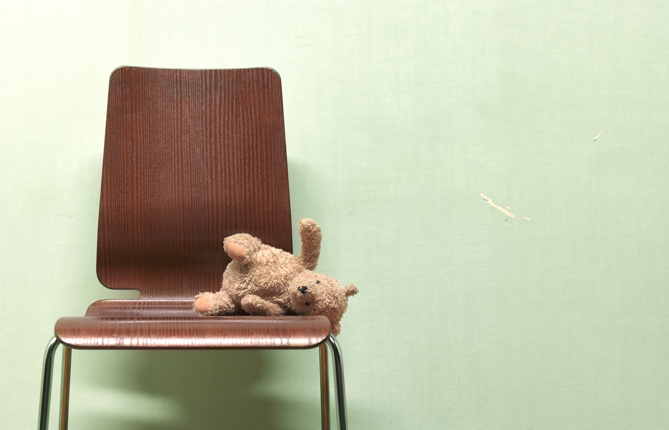 A wooden chair is against a plain wall with a stuffed teddy bear lying on its side on the chair