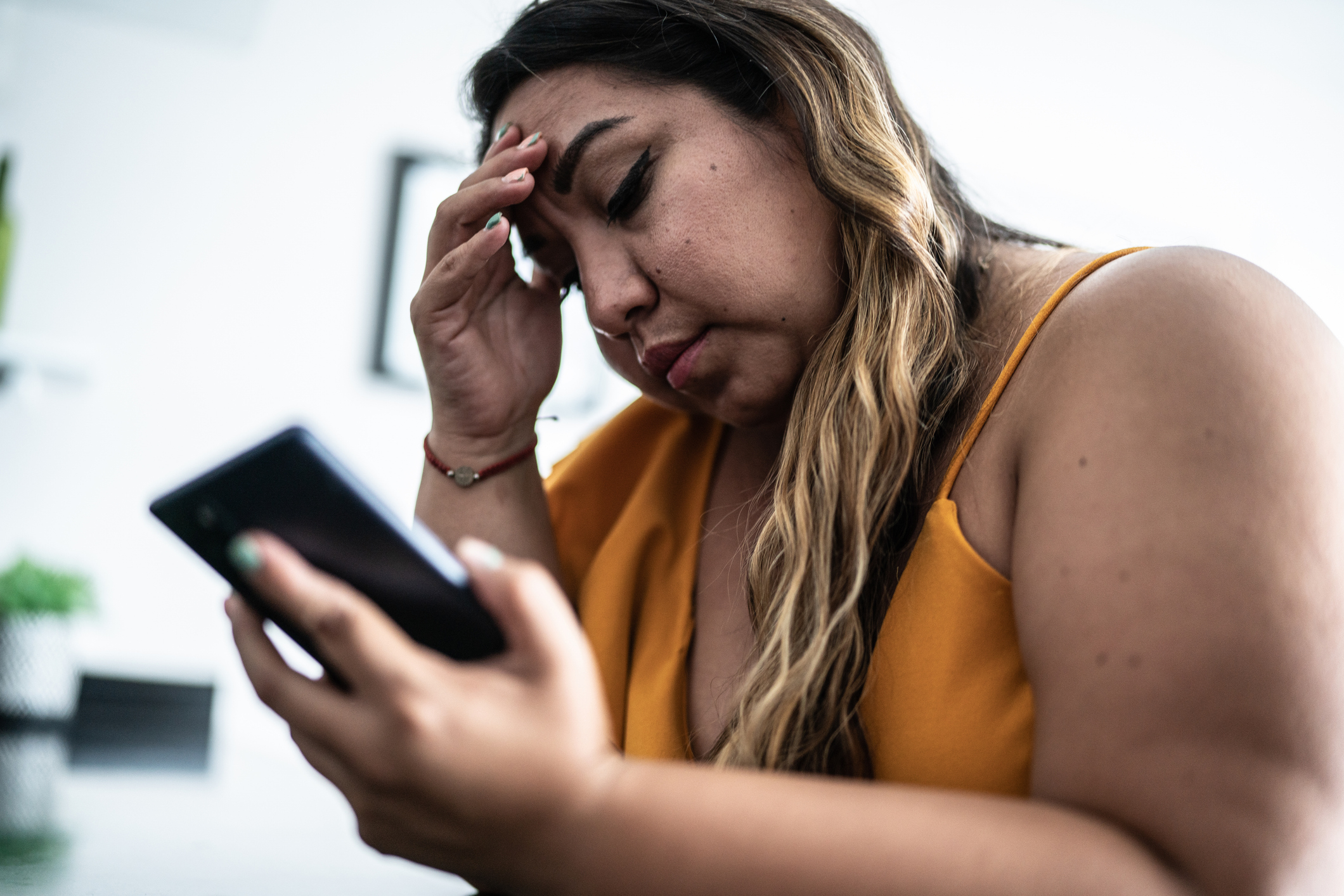 Woman looks stressed, holding her head with one hand and a smartphone in the other, sitting indoors