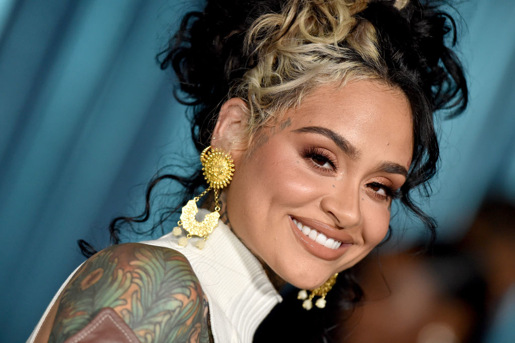 Kehlani smiles at an event, wearing a sleeveless turtleneck, large gold earrings, and sporting a detailed arm tattoo