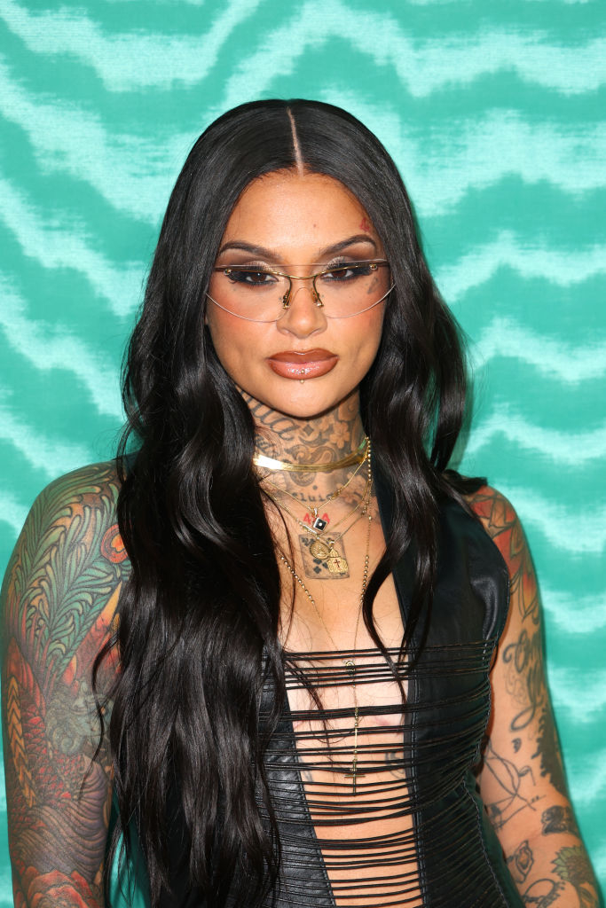Kehlani poses in front of a patterned backdrop, wearing a black cut-out dress, layered necklaces, tinted glasses, and showcasing full-arm tattoos