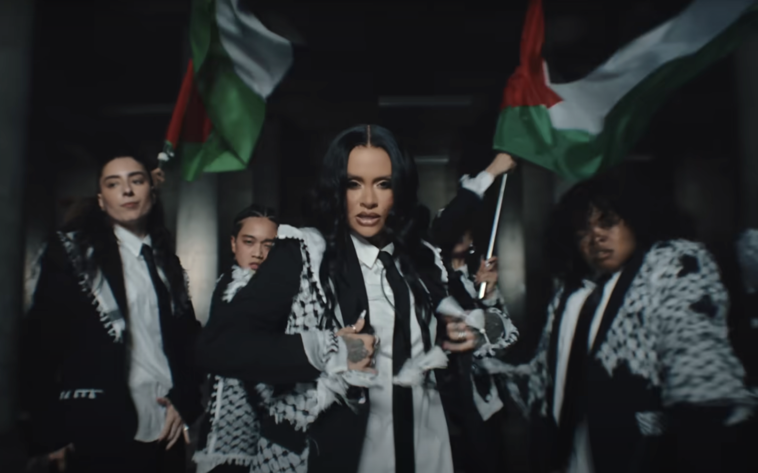 Group of people, including singer Kehlani, wearing patterned outfits and holding flags while posing confidently in what appears to be a music video scene