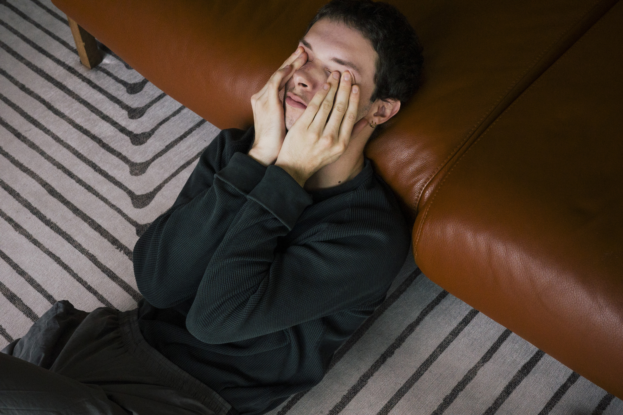 A person wearing a sweater sits on the floor with hands covering the face, leaning against a brown sofa on a striped carpet