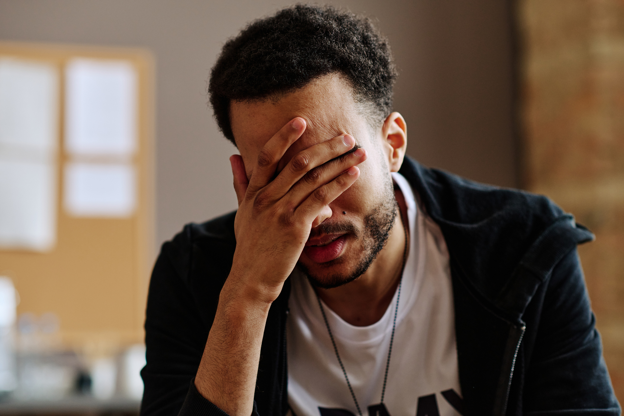 A person covering their face with one hand, appearing stressed or frustrated. A bulletin board is visible in the background