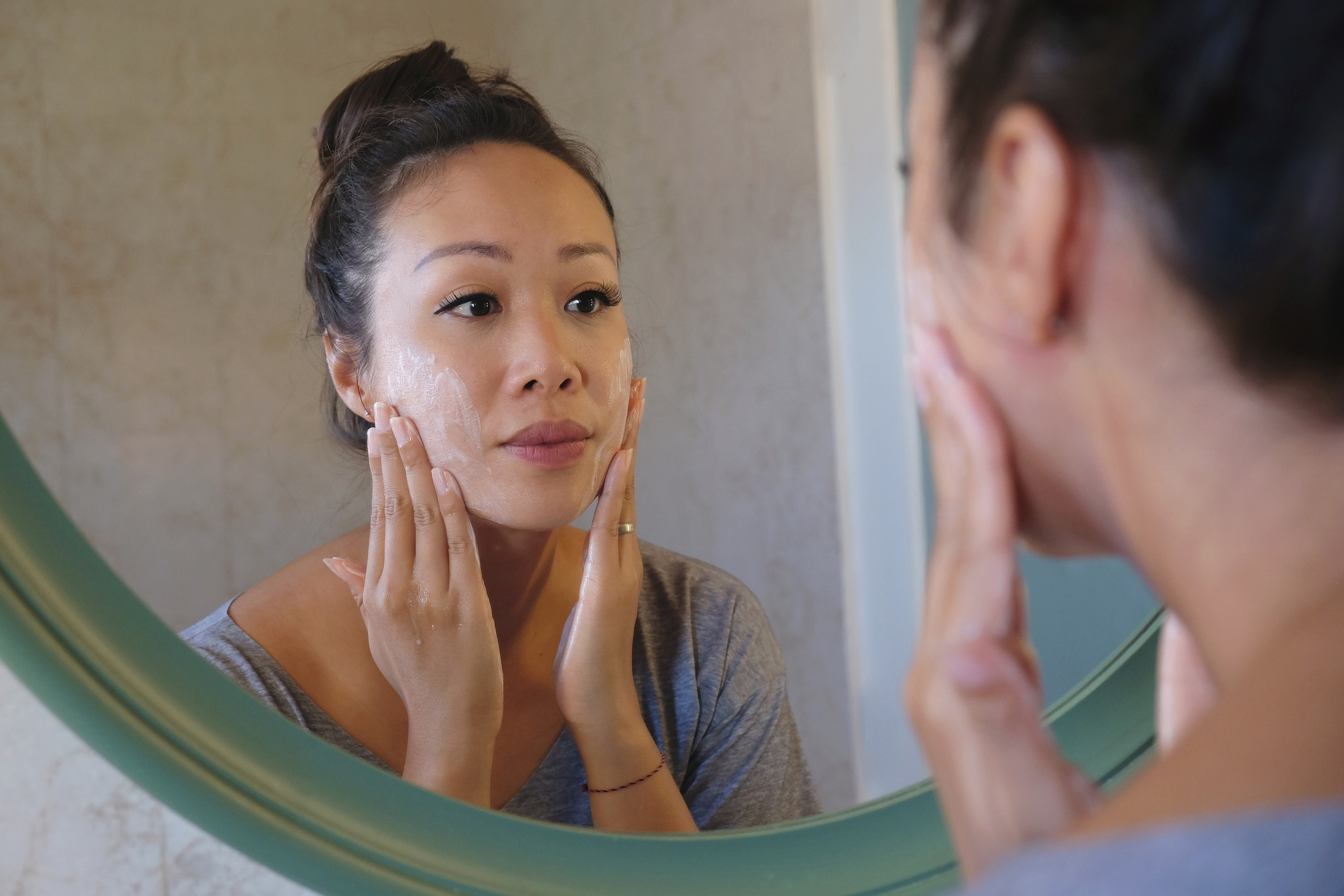 A woman is cleansing her face in front of a mirror, gently massaging her cheeks with both hands