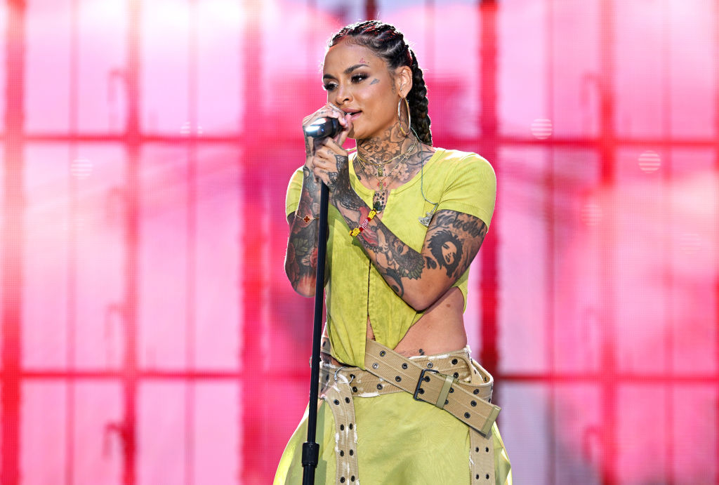 Kehlani performing on stage, wearing a stylish outfit featuring intricate tattoos, a short-sleeve top, and a decorative belt, holding a microphone