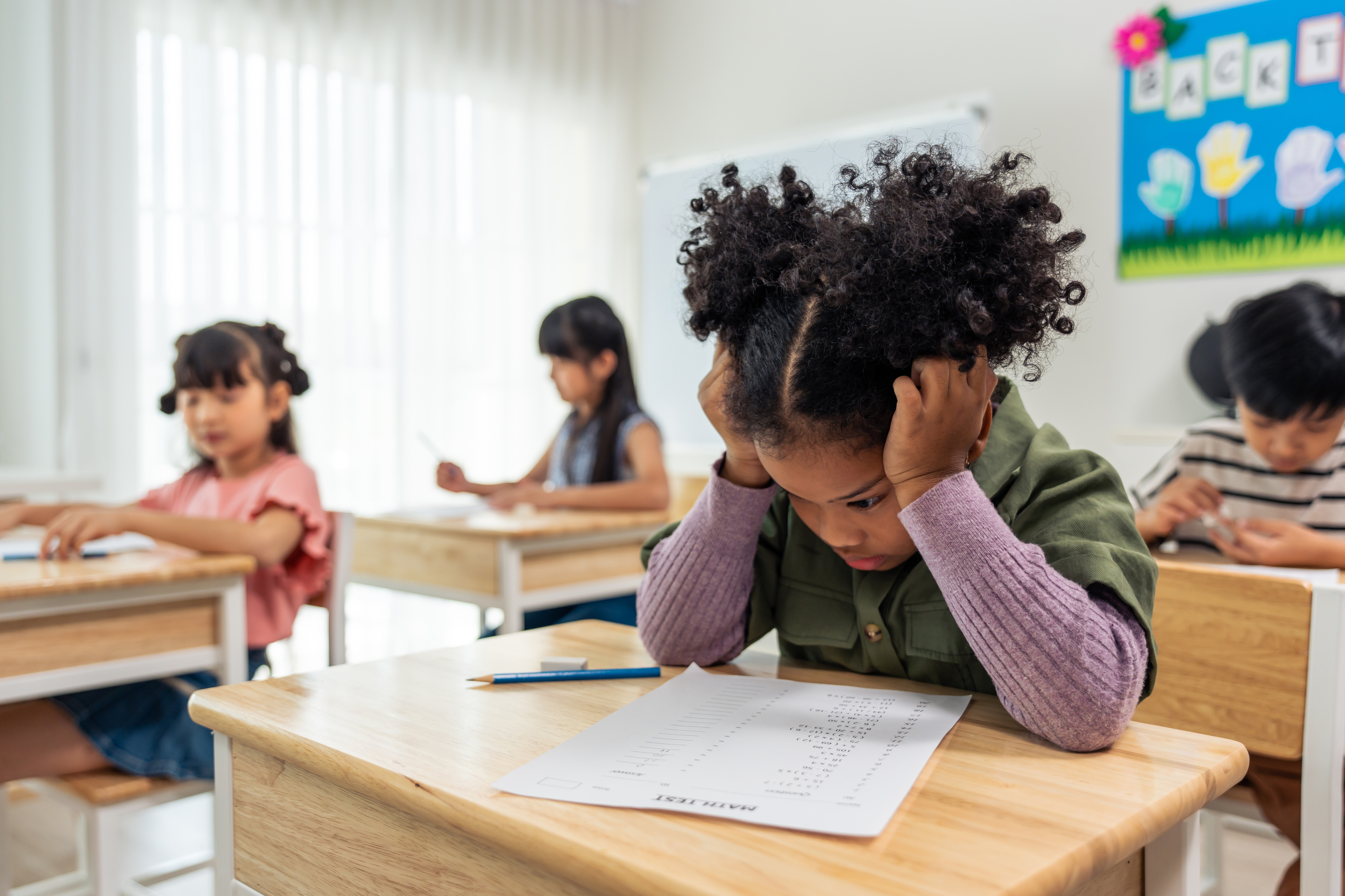 Children focus on their schoolwork at desks in a classroom. A girl in the foreground appears frustrated, with her head in hands