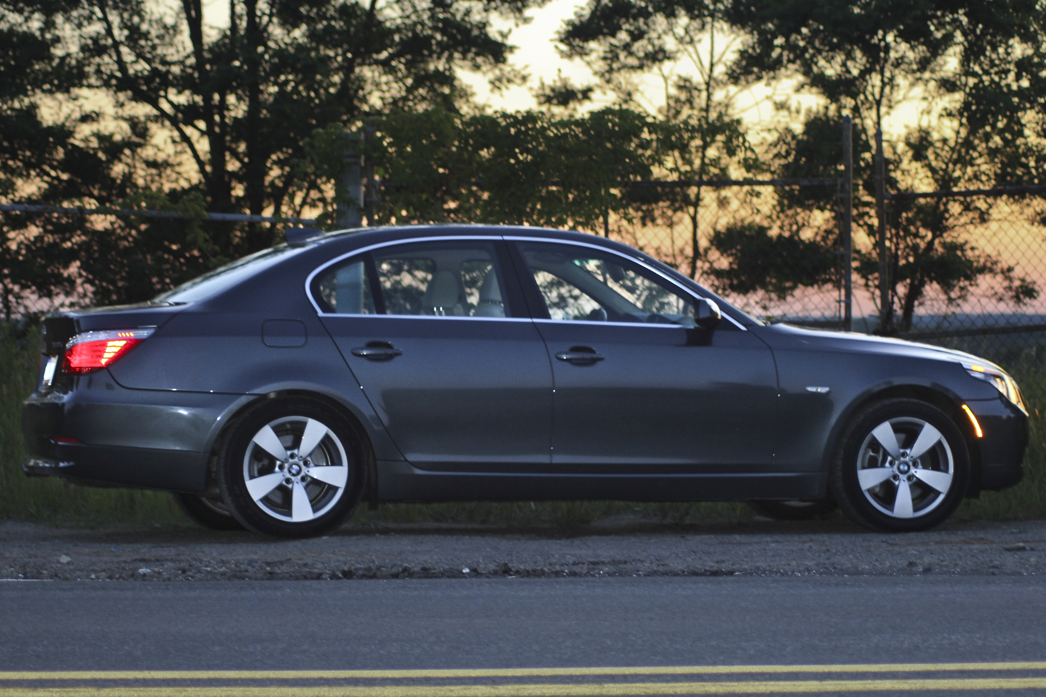 A side view of a dark-colored sedan parked on a roadside with trees and a sunset in the background