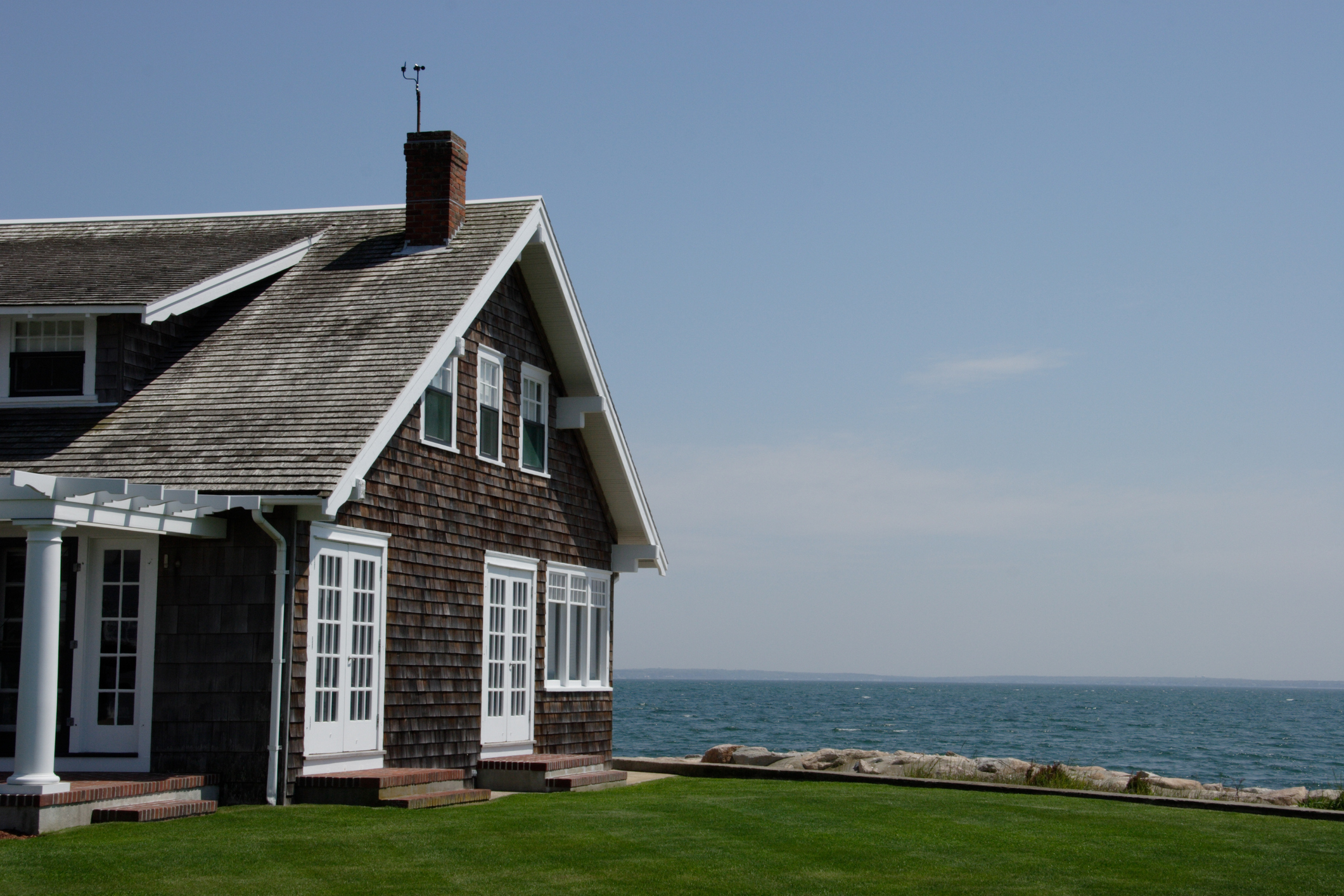 A coastal house with wooden shingles, large windows, and a chimney overlooks the ocean on a sunny day