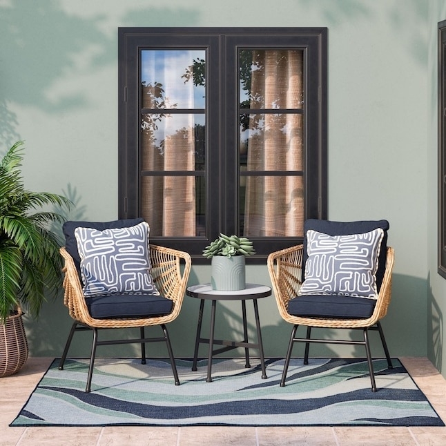Two rattan chairs with patterned cushions face a small round table holding a potted plant on an outdoor patio with a stylish wave-pattern rug