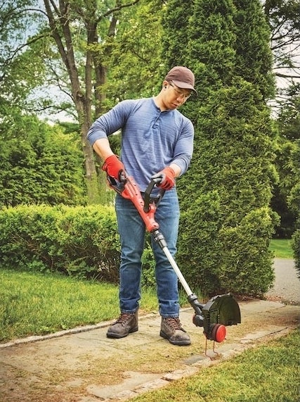 A person wearing a hat, glasses, long-sleeved shirt, jeans, and gloves is using an electric trimmer to edge a lawn in a lush, green garden area