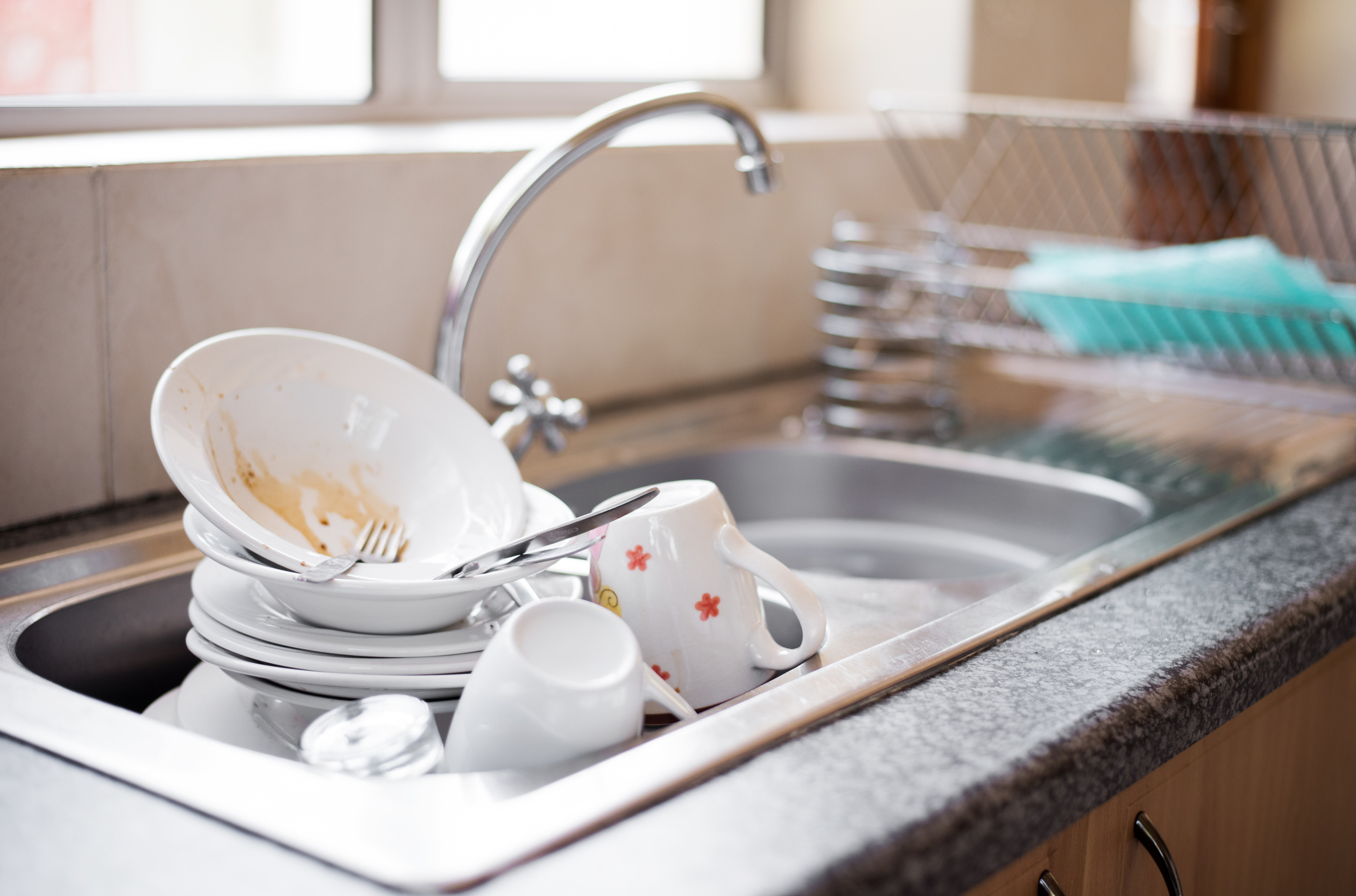 A kitchen sink filled with dirty dishes, including plates, bowls, mugs, and cutlery, sitting atop the counter next to a drying rack