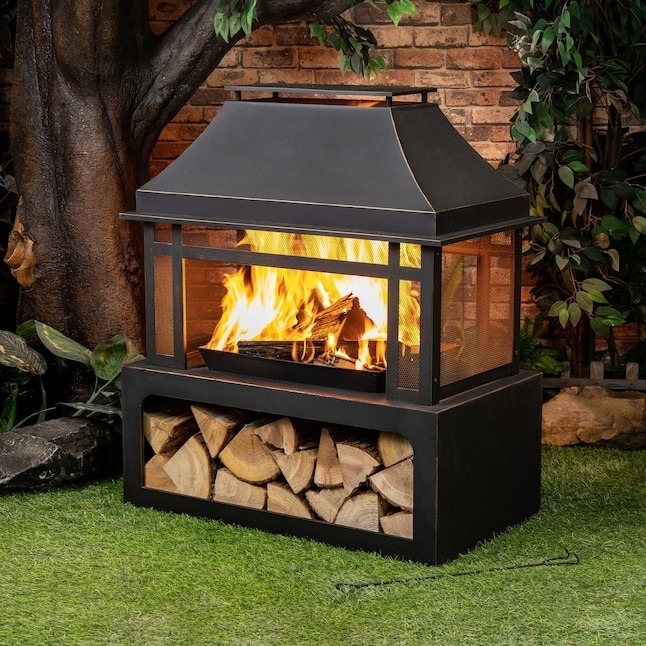 A black outdoor fireplace with logs burning inside. A storage area below holds extra firewood. The setup is in a garden with brick and tree background