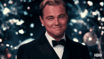 Leonardo DiCaprio is smiling in a formal tuxedo with festive lights in the background