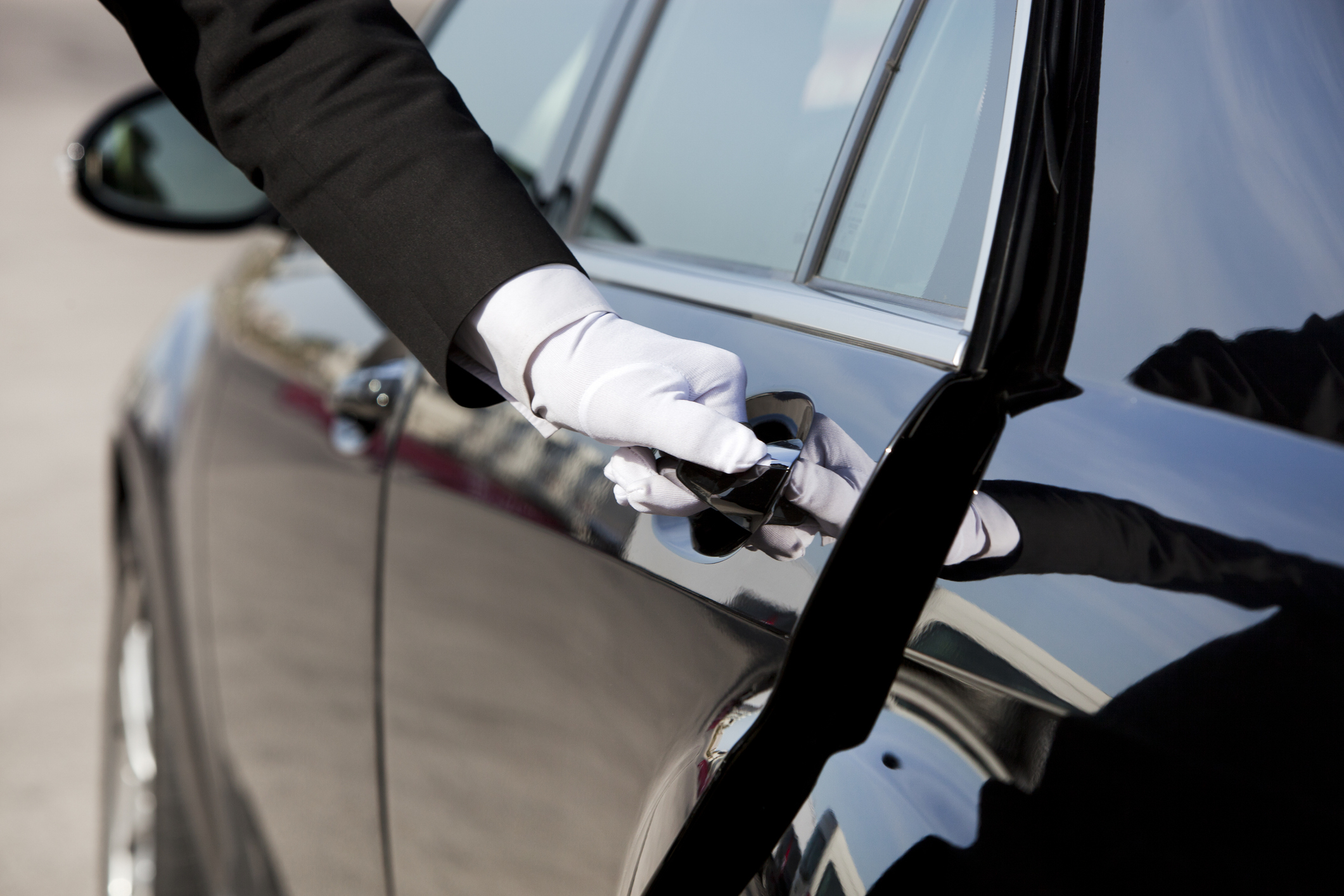 A chauffeur dressed in a black suit and white gloves opens the door of a luxury car