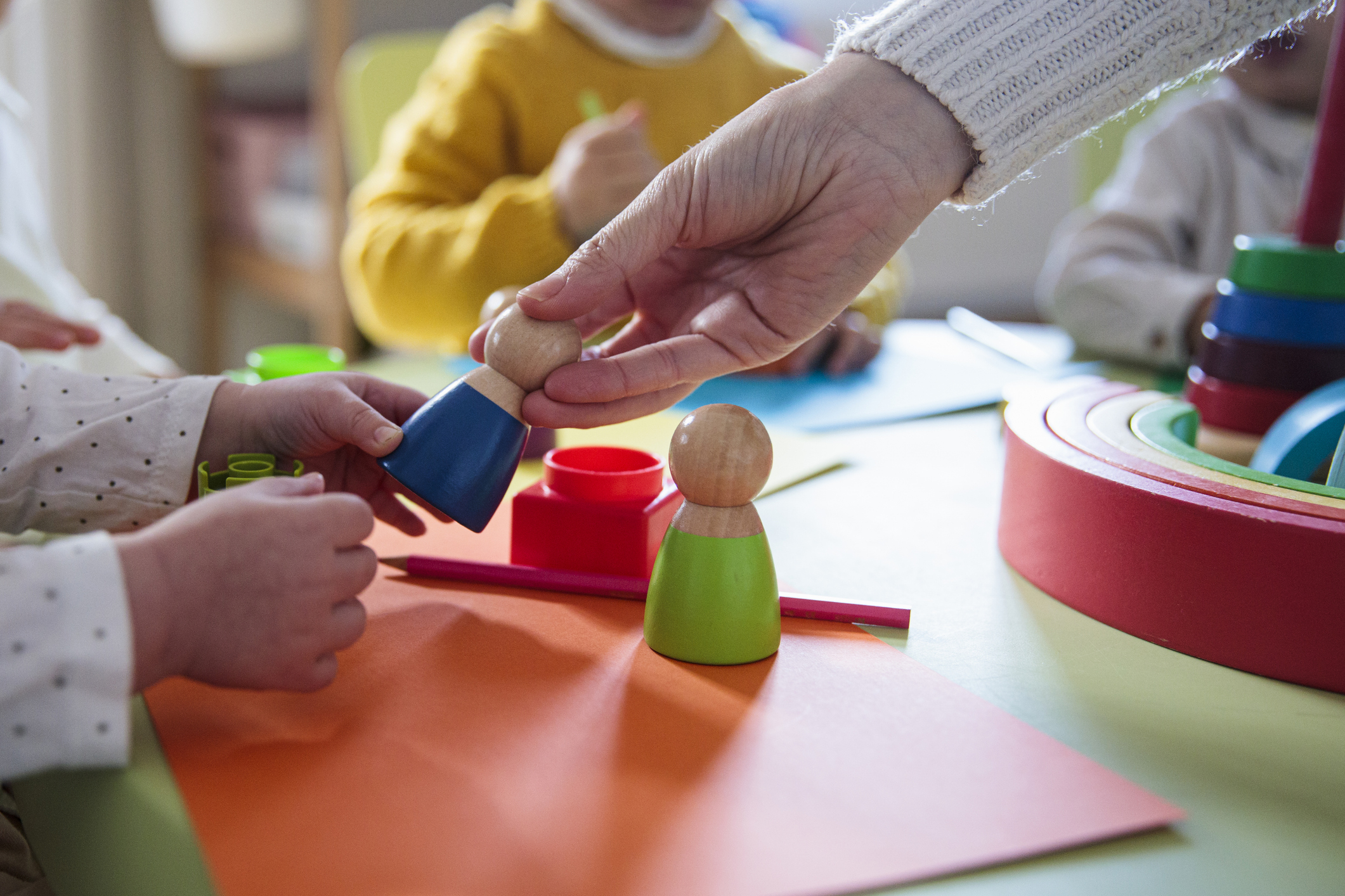 A close-up of children playing with wooden peg dolls and other toys at a table, with an adult handing a peg doll to one of the children