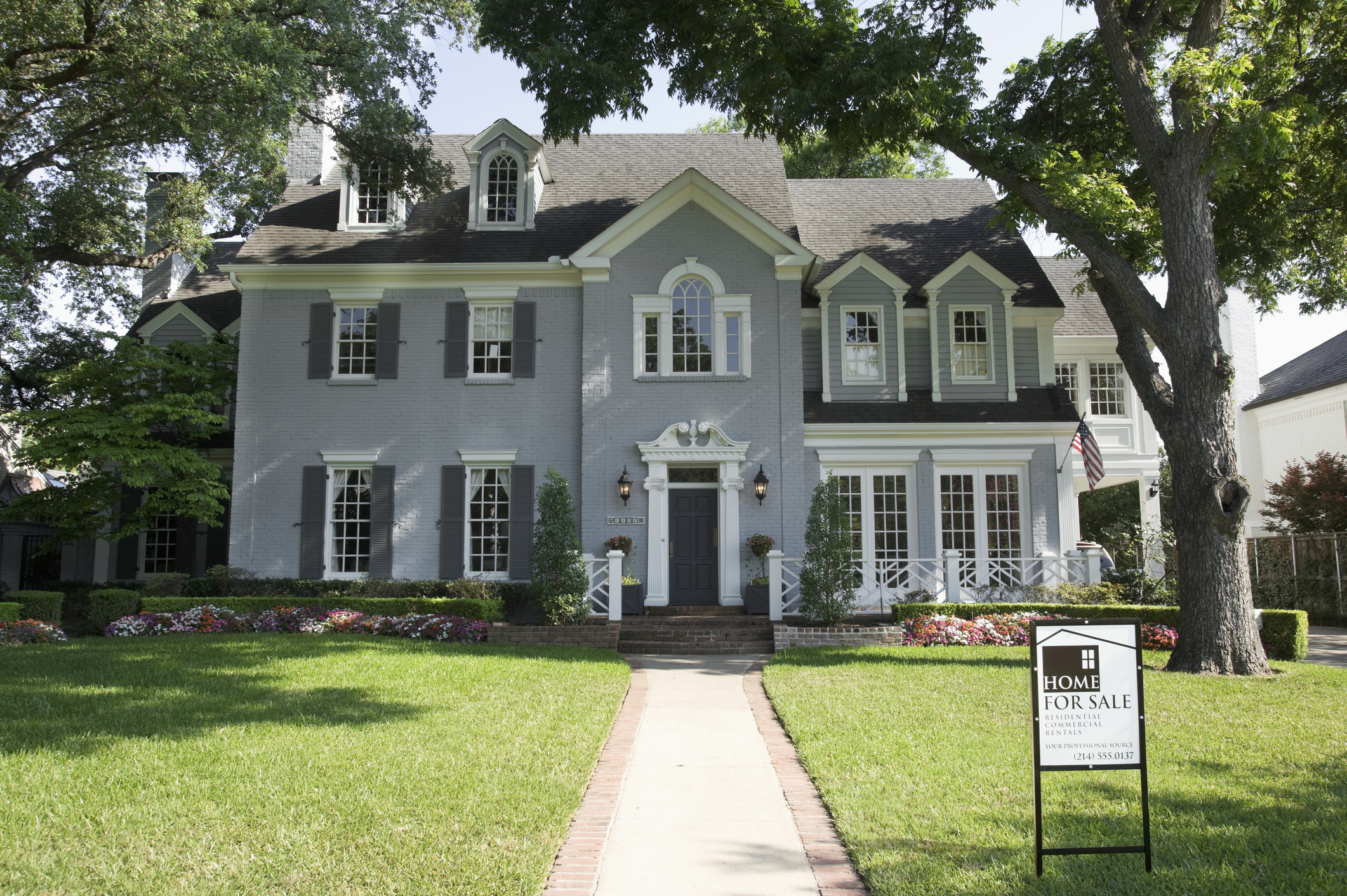 A large two-story house with a &quot;Home for Sale&quot; sign in the front yard, surrounded by a well-manicured lawn and shaded by trees