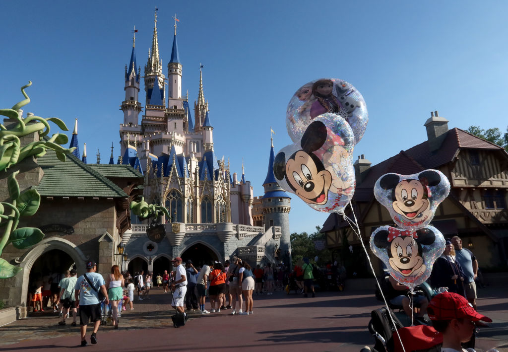 Disney theme park with a castle in the background and Mickey Mouse balloons in the foreground. Visitors are walking and standing in front of the castle