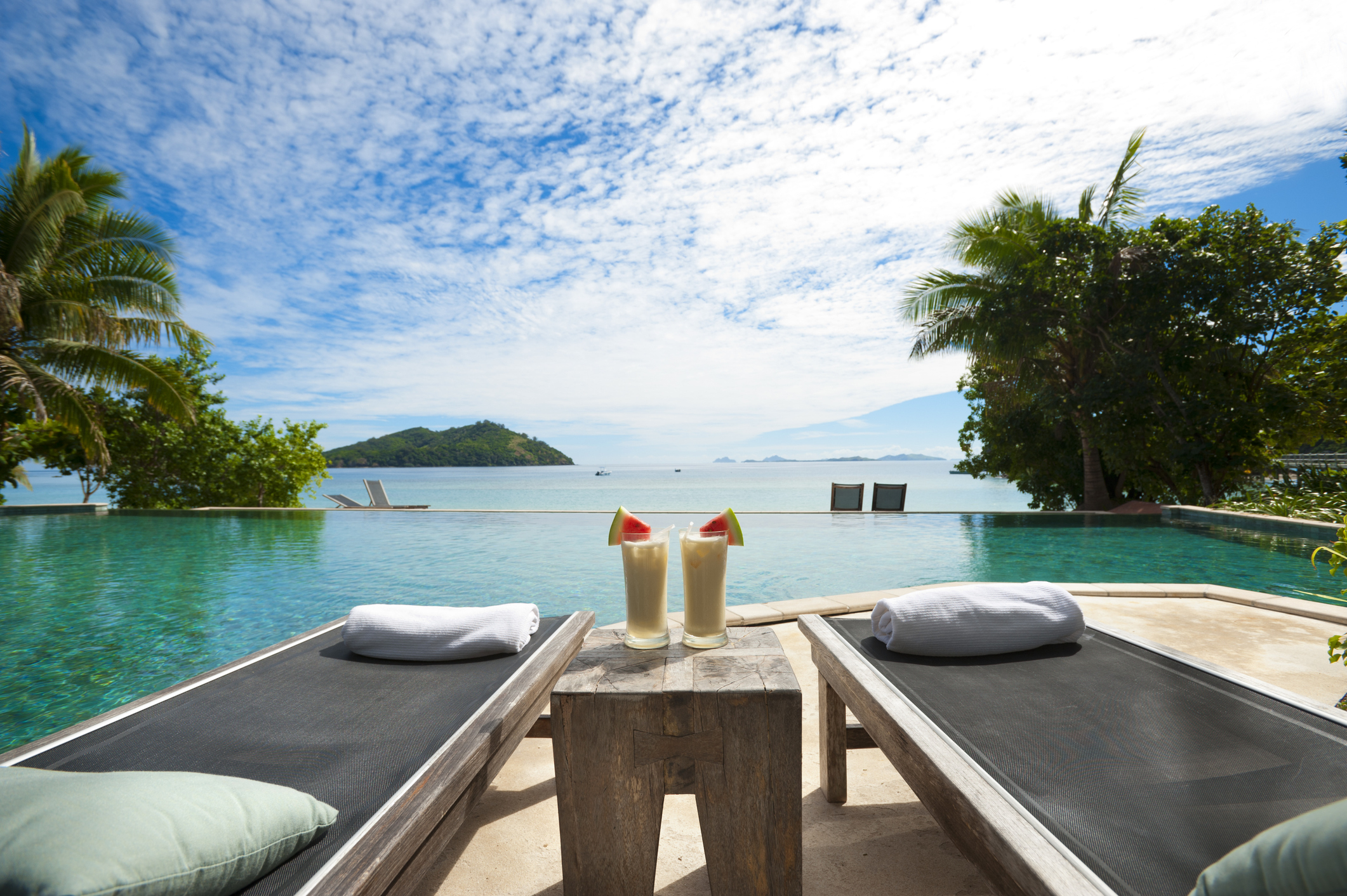 Two lounge chairs facing a scenic infinity pool with two tropical drinks on a small table between them. Background includes lush greenery and a distant island