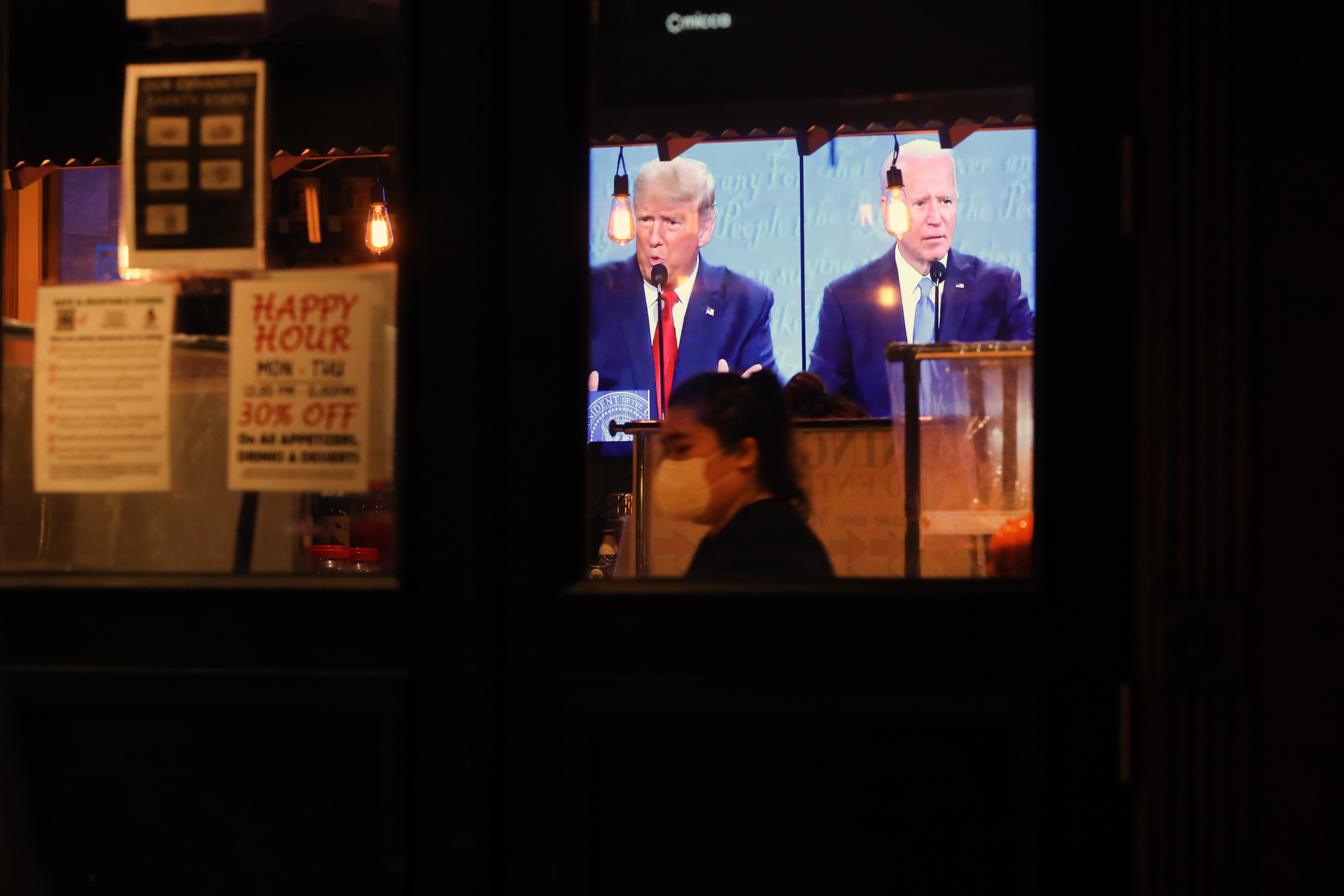 Donald Trump and Joe Biden appear on a TV screen inside a dimly lit restaurant, as a person in a mask stands in the foreground