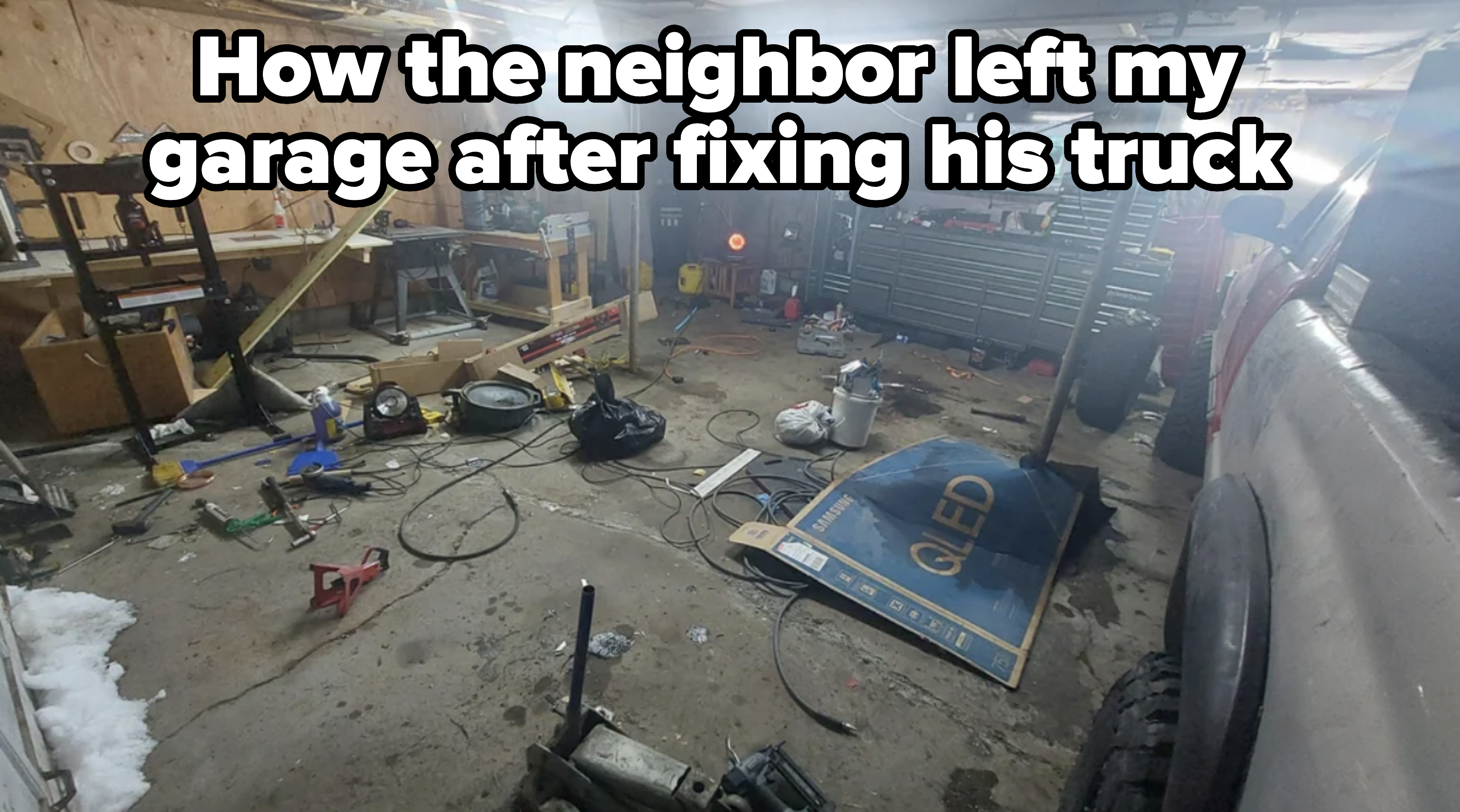 A cluttered garage with various tools, tires, and equipment scattered around. There is a long workbench against one wall and a large toolbox on the other side