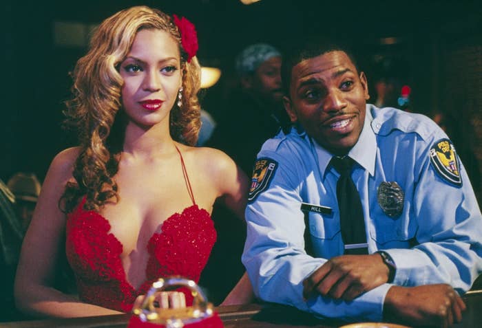 Beyoncé in a glamorous dress with a flower accessory sits next to Mekhi Phifer in a police uniform at a casino-like setting