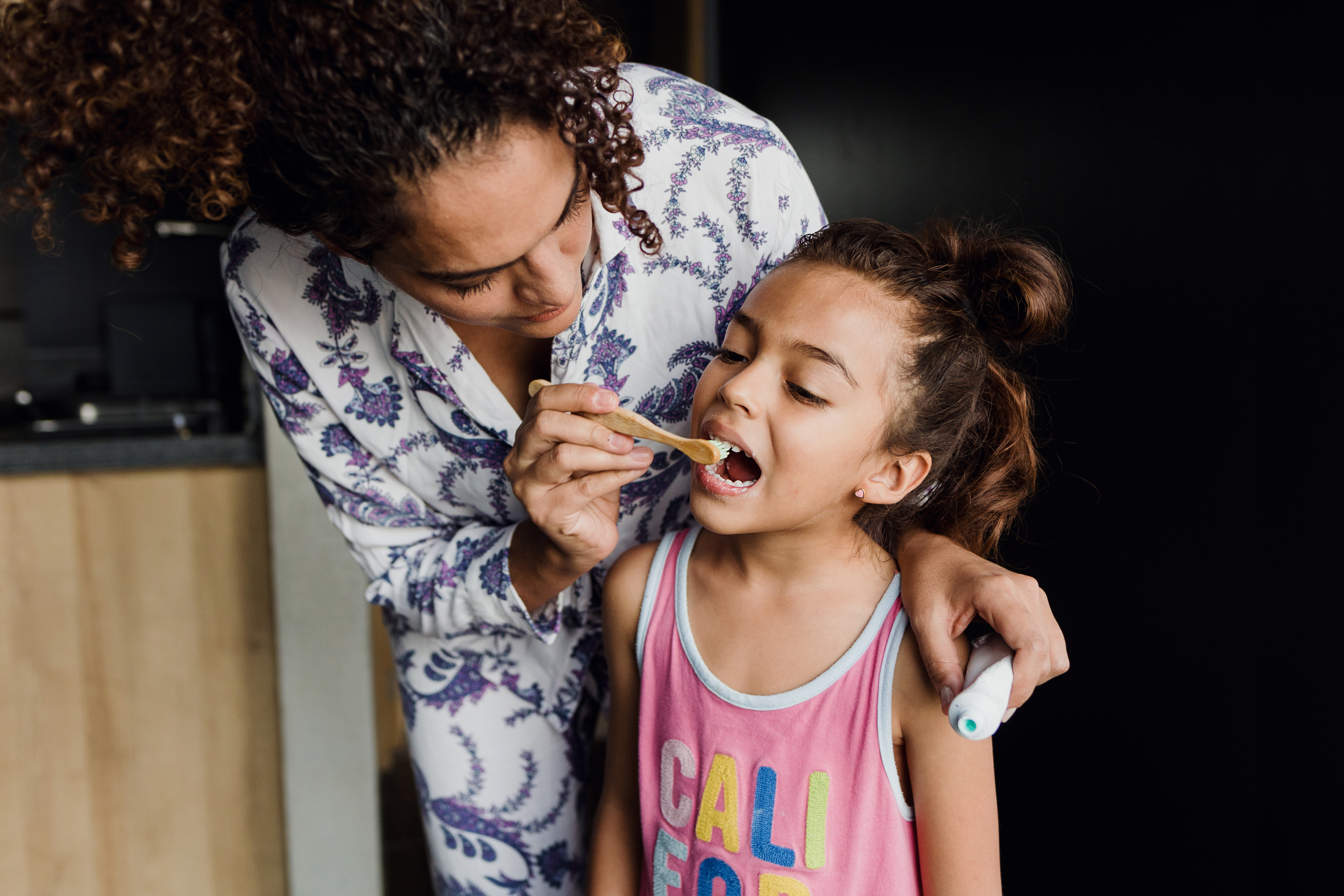 A woman in pajamas helps a young girl in a sleeveless shirt brush her teeth