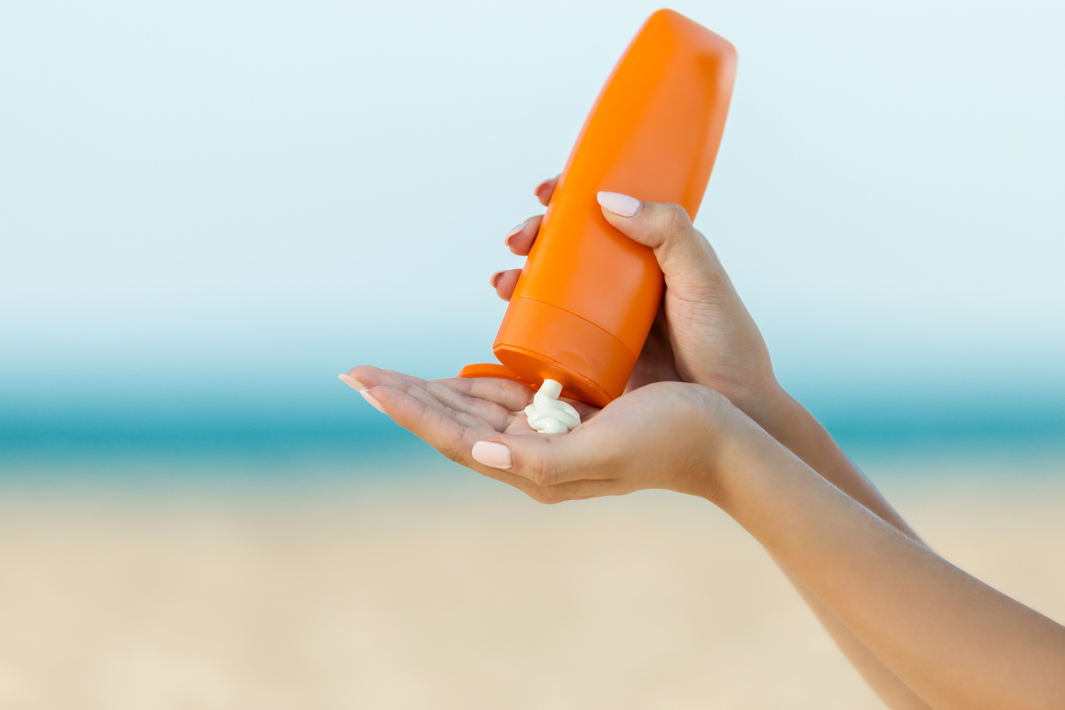 A person is squeezing sunscreen from an orange bottle into their hand on a sandy beach