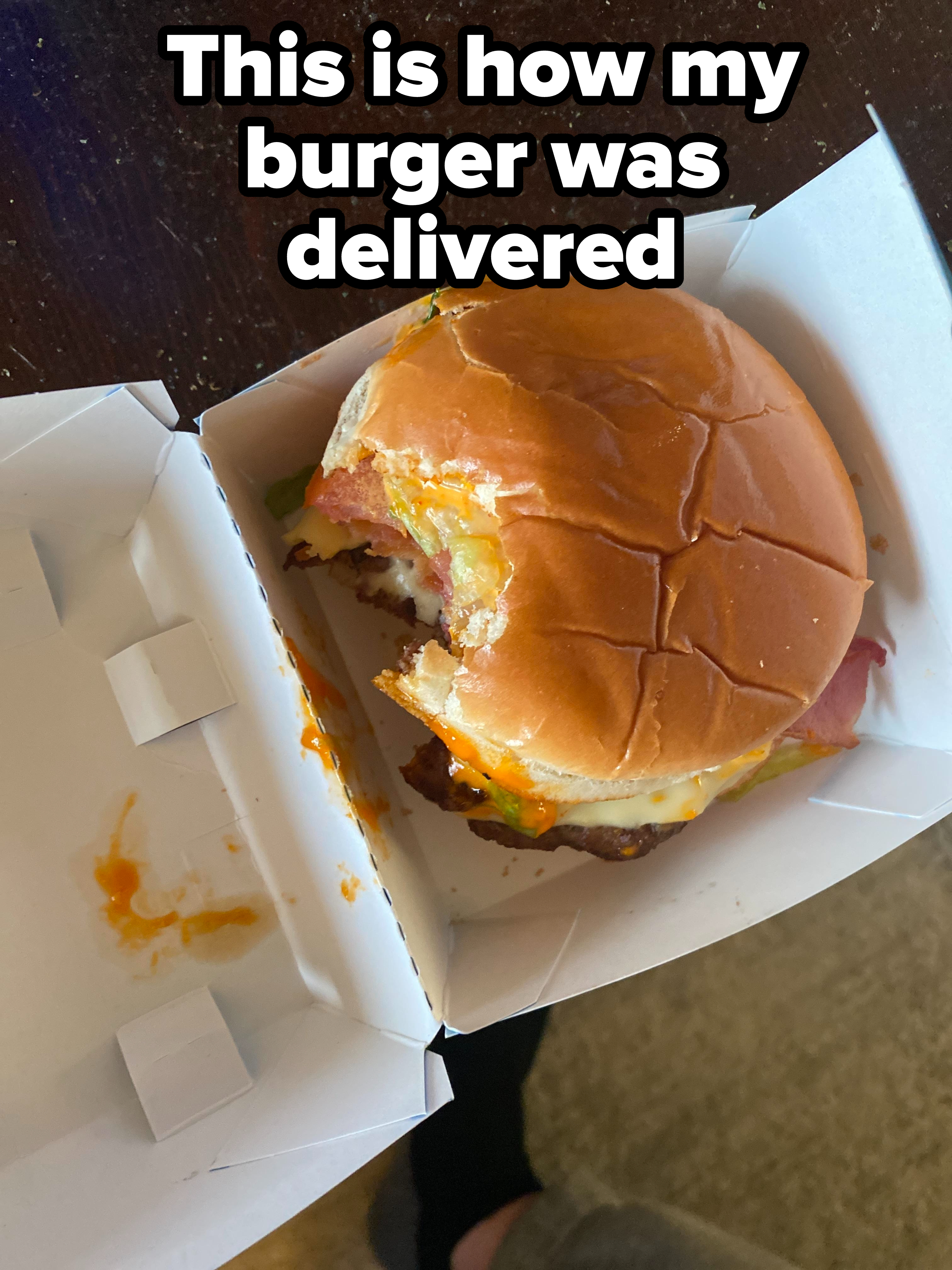 Partially eaten burger with visible condiments and fillings, in an open takeout box on a wooden surface