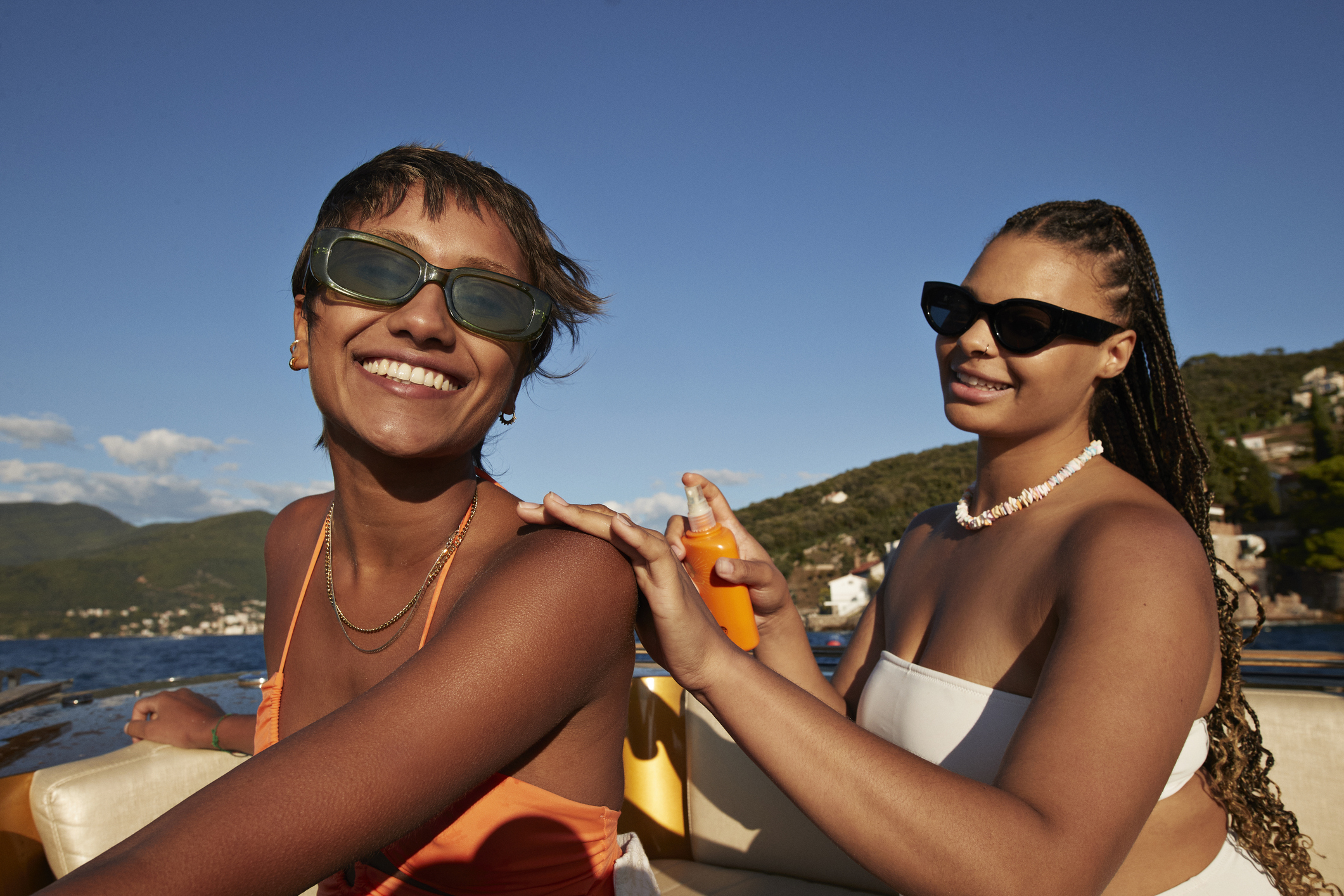 Two women, one with short hair and sunglasses, the other with braids and sunglasses, enjoying a boat ride while applying sunscreen