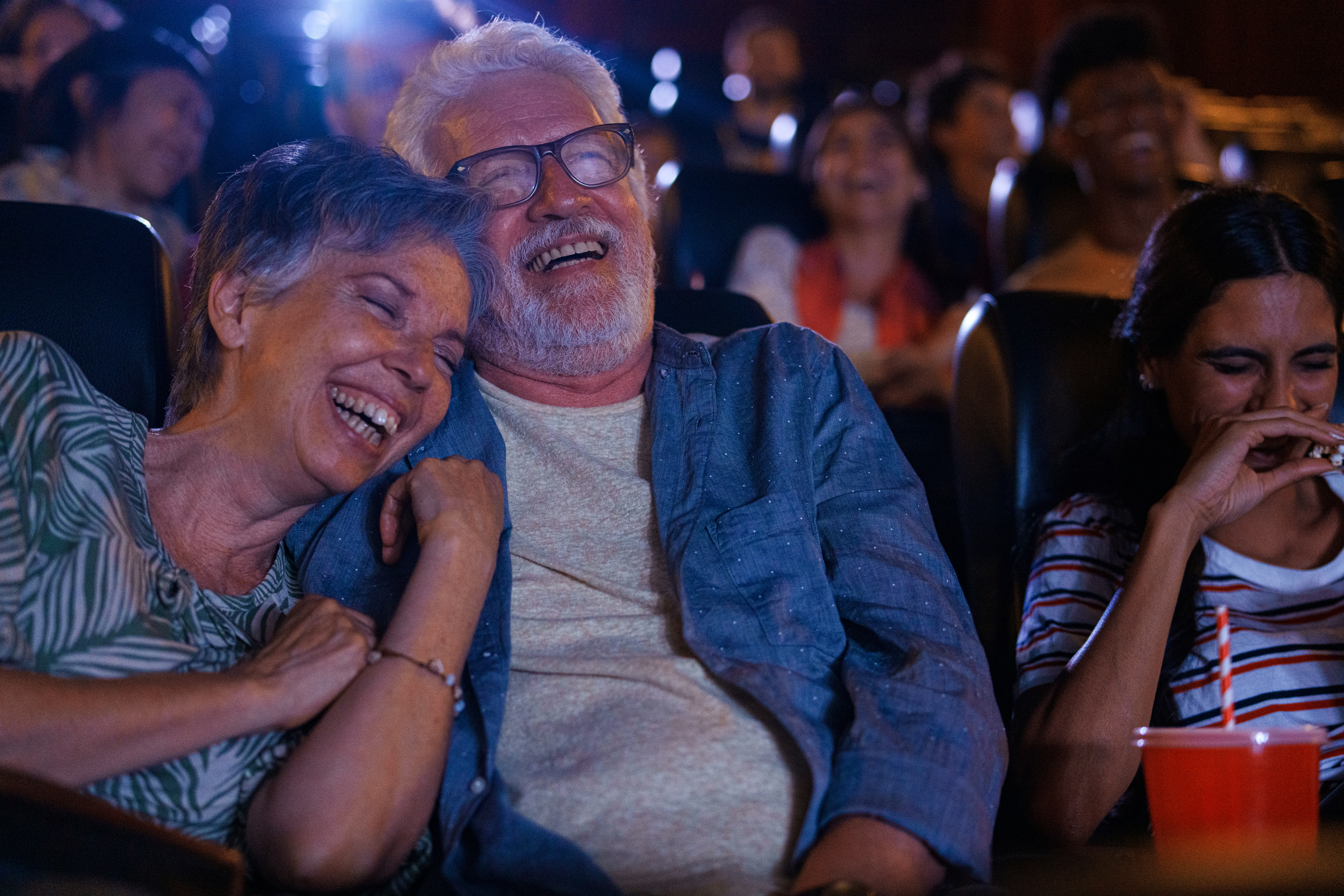 An elderly couple and a woman laugh while watching a movie in a theater, surrounded by other smiling people