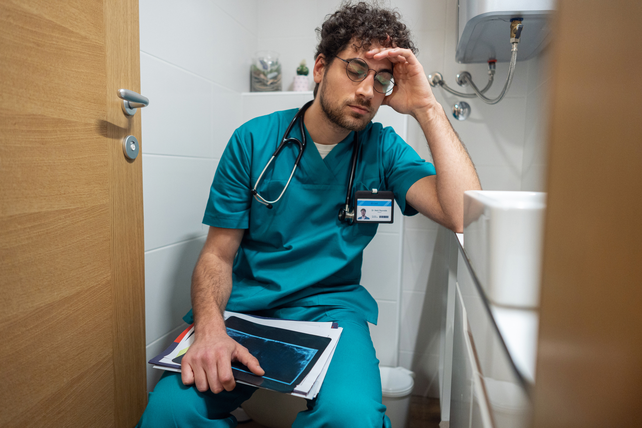 A tired doctor in scrubs holding medical files sits with his head resting on his hand in a small room, which appears to be a restroom