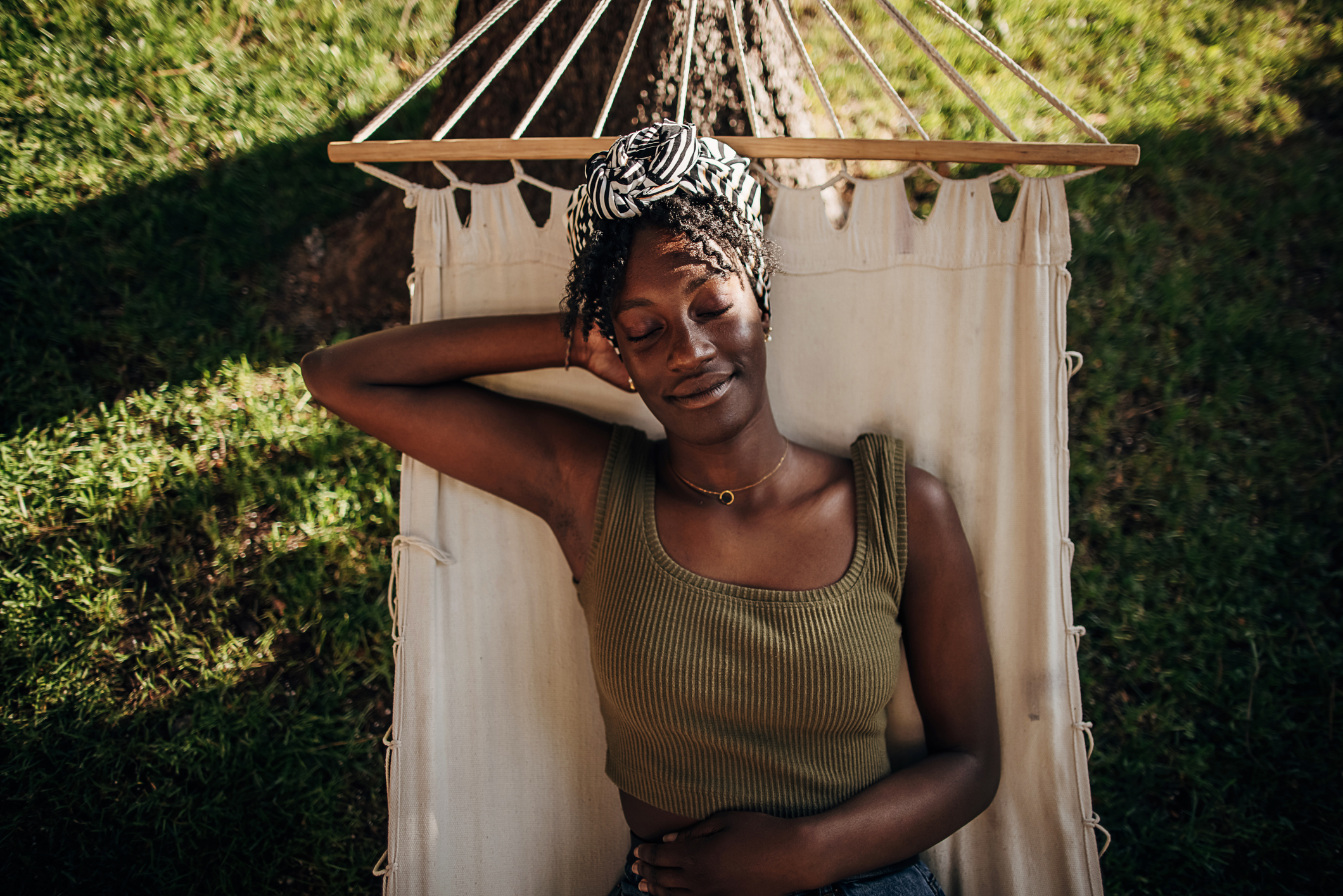A person relaxes with eyes closed in a hammock under a tree, enjoying a peaceful moment outdoors. They wear a sleeveless top and a head wrap