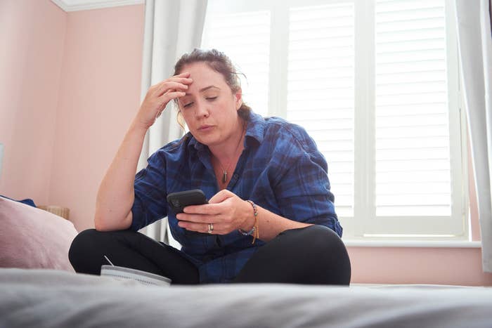 A woman sits on a bed, looking stressed while reading something on her phone