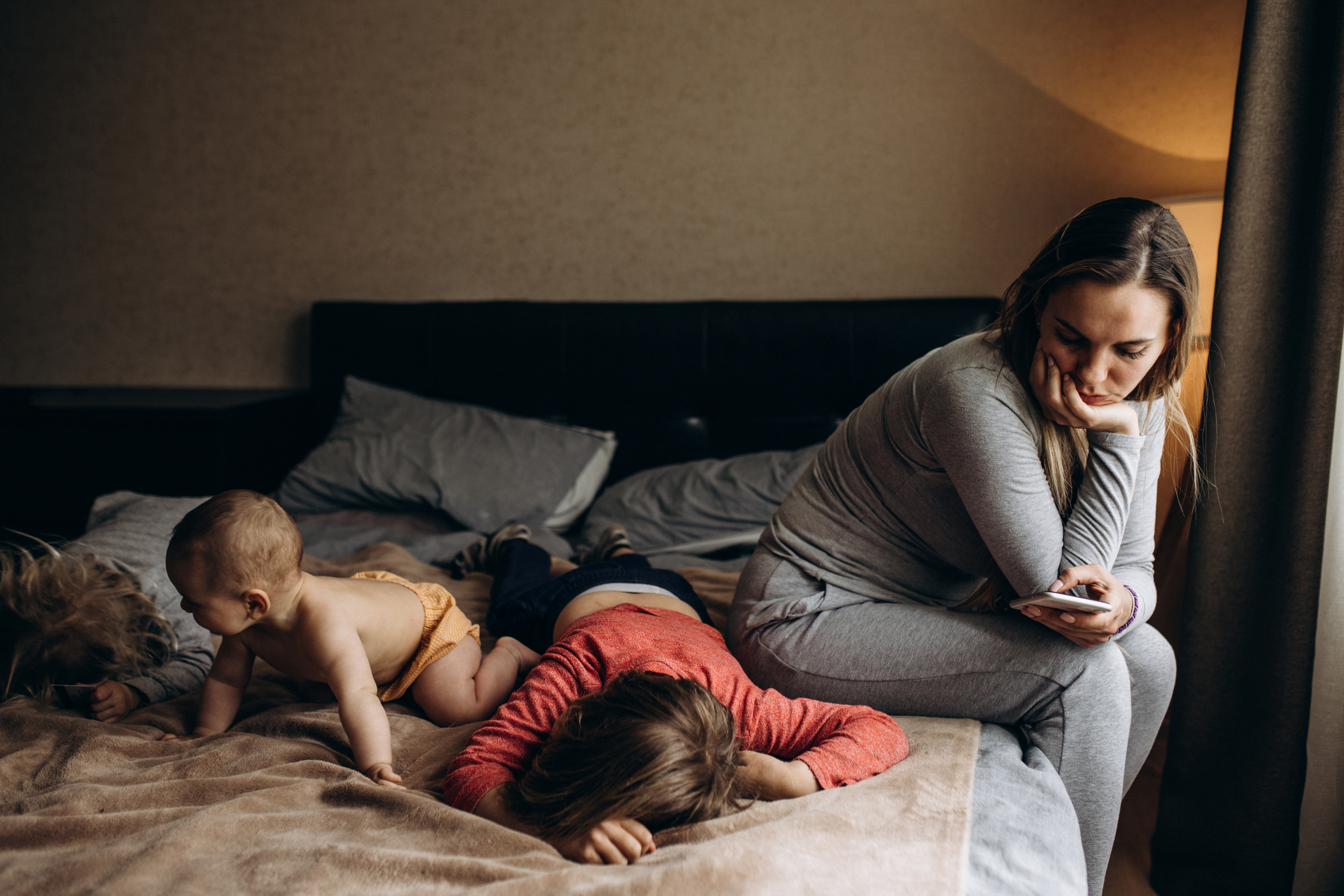A woman in casual clothing looks at her phone while sitting on a bed. Two young children, one crawling and one lying down, are on the bed beside her