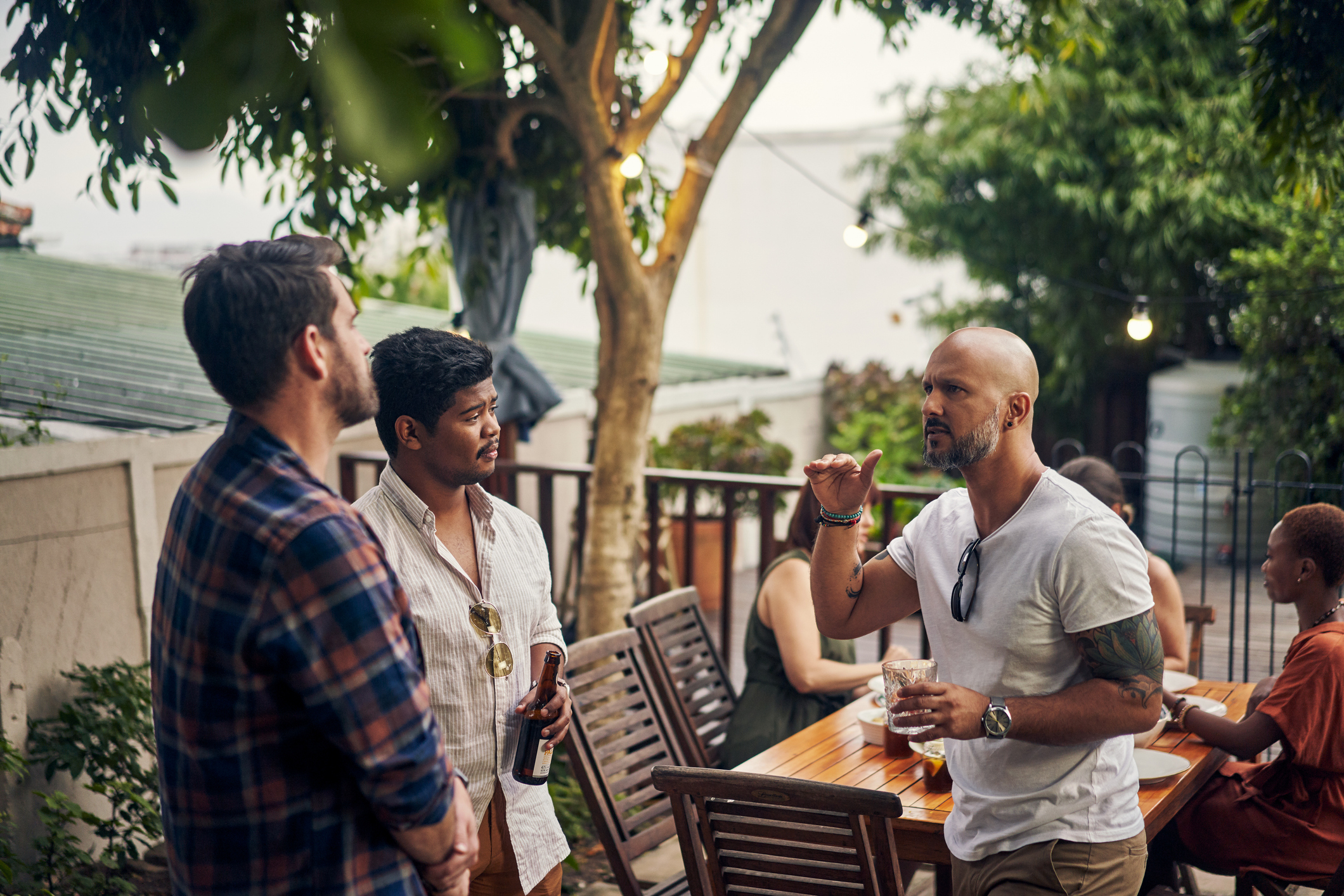 Group of people enjoying an outdoor gathering; three men in the foreground engaging in conversation, with others seated around a wooden table in the background