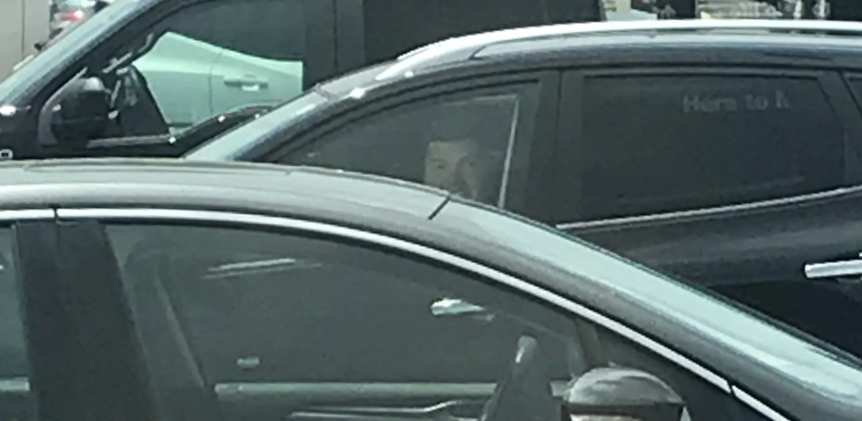 A person is seen sitting inside a car, partially hidden by the car window and frame. The expression is not clearly visible