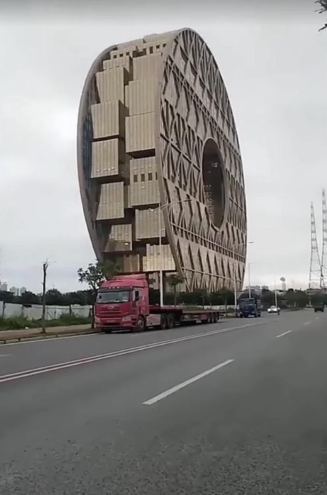 A large, round building with unique rectangular windows and geometric design stands beside a road with vehicles passing by