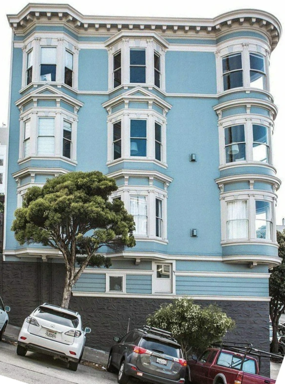 Four-story building with ornate architectural details, featuring multiple tall windows. There are several cars parked in front and a tree partially obscuring part of the building