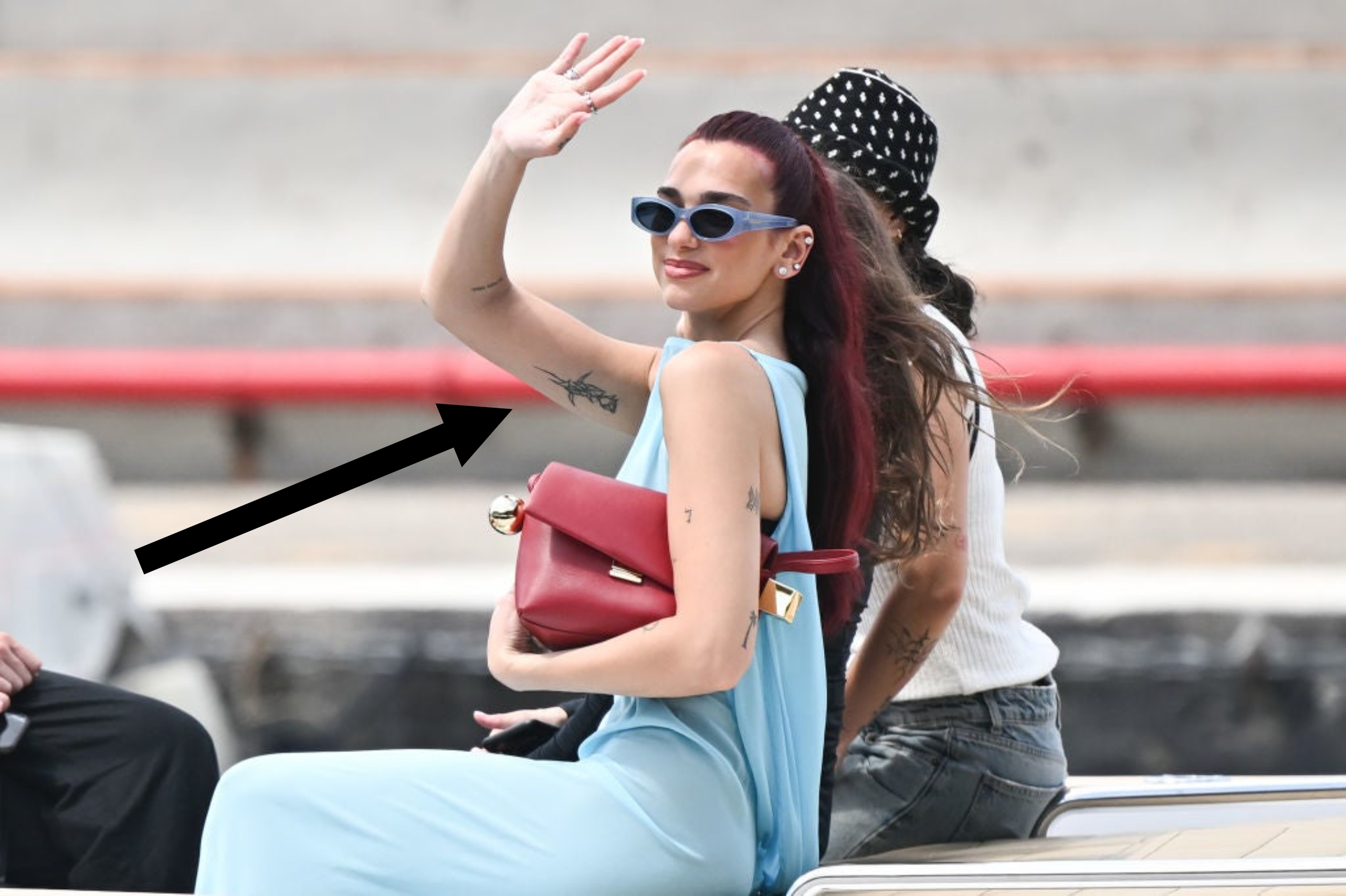 Dua Lipa, with a tattooed arm, in a stylish sleeveless outfit, blue sunglasses, and carrying a red handbag, waves while seated outdoors beside another individual