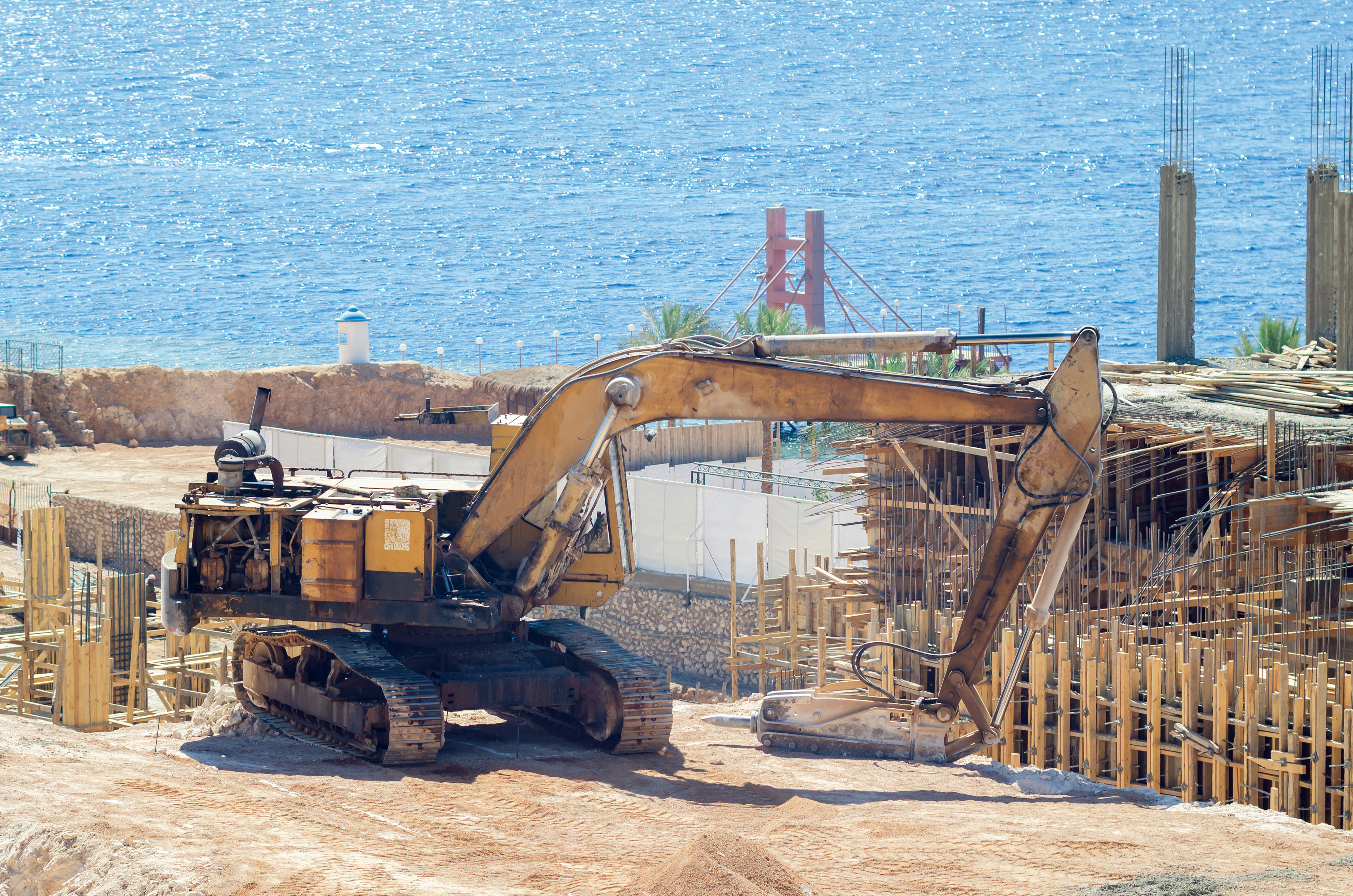 An excavator working at a construction site near the coast, with the sea and construction materials in the background