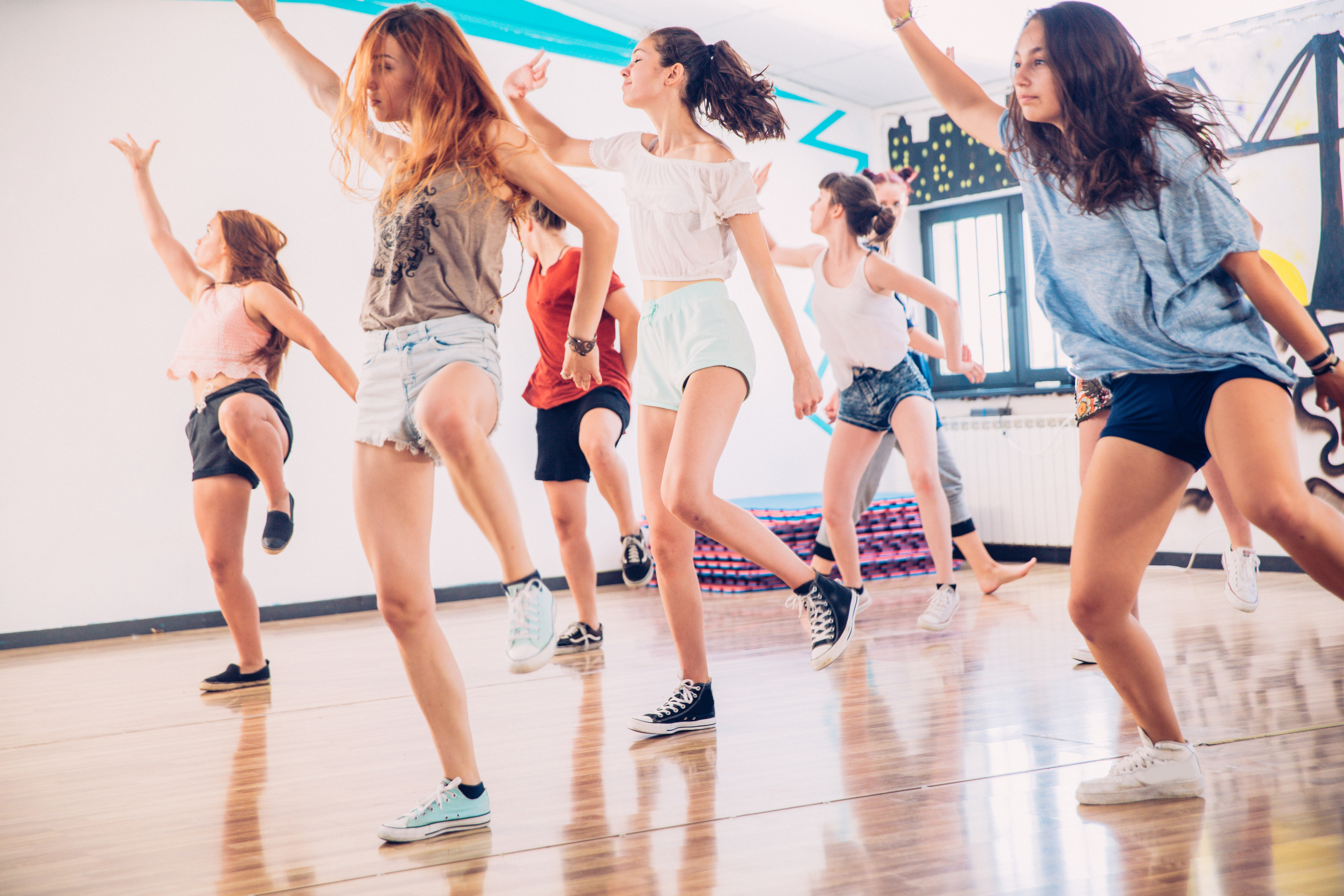 A group of young girls  in casual clothing practice a dance routine in a studio with mirrors and wooden floors.