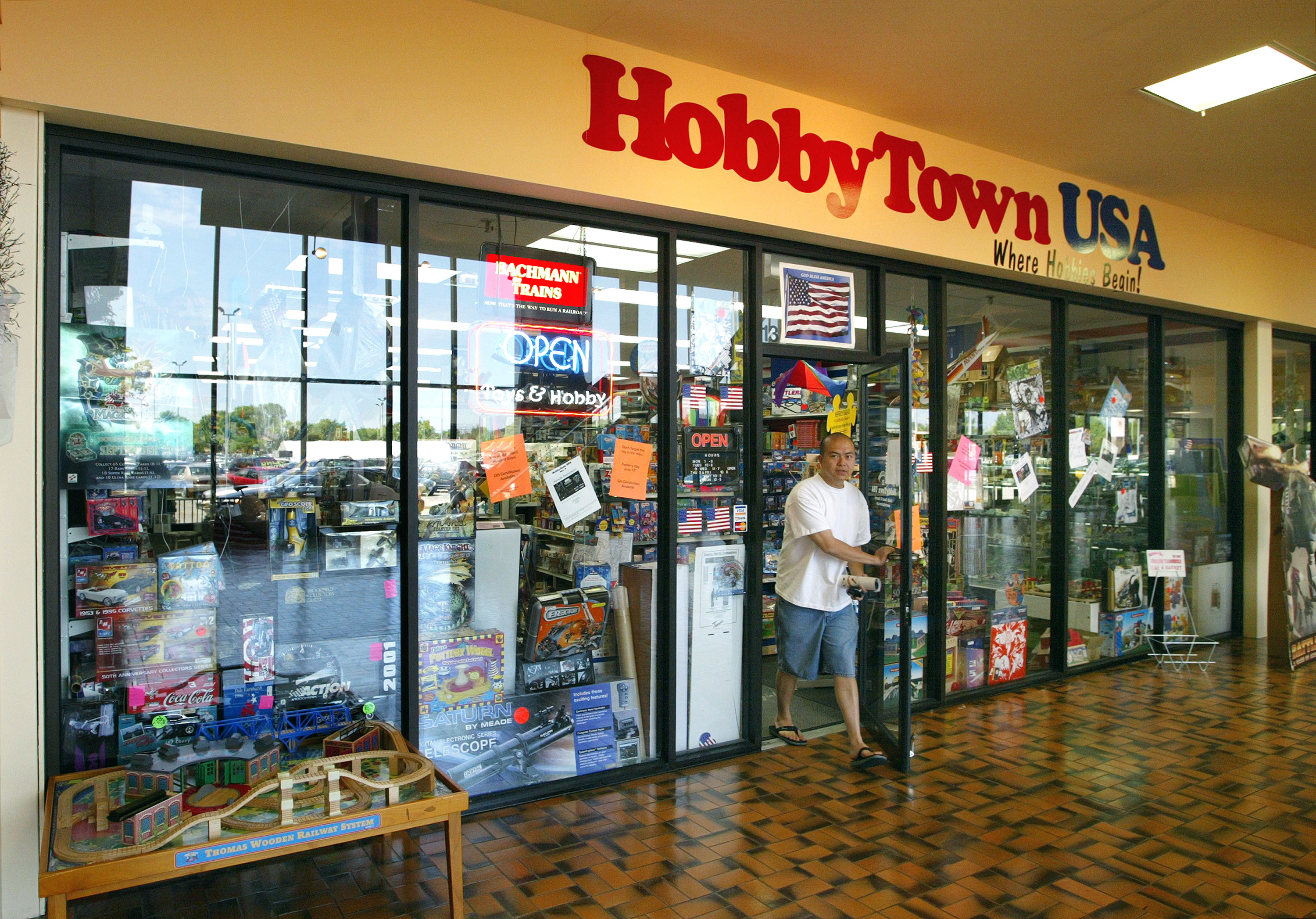 A man exits a HobbyTown USA store while holding a bag. The storefront features various hobby-related items on display and several signs