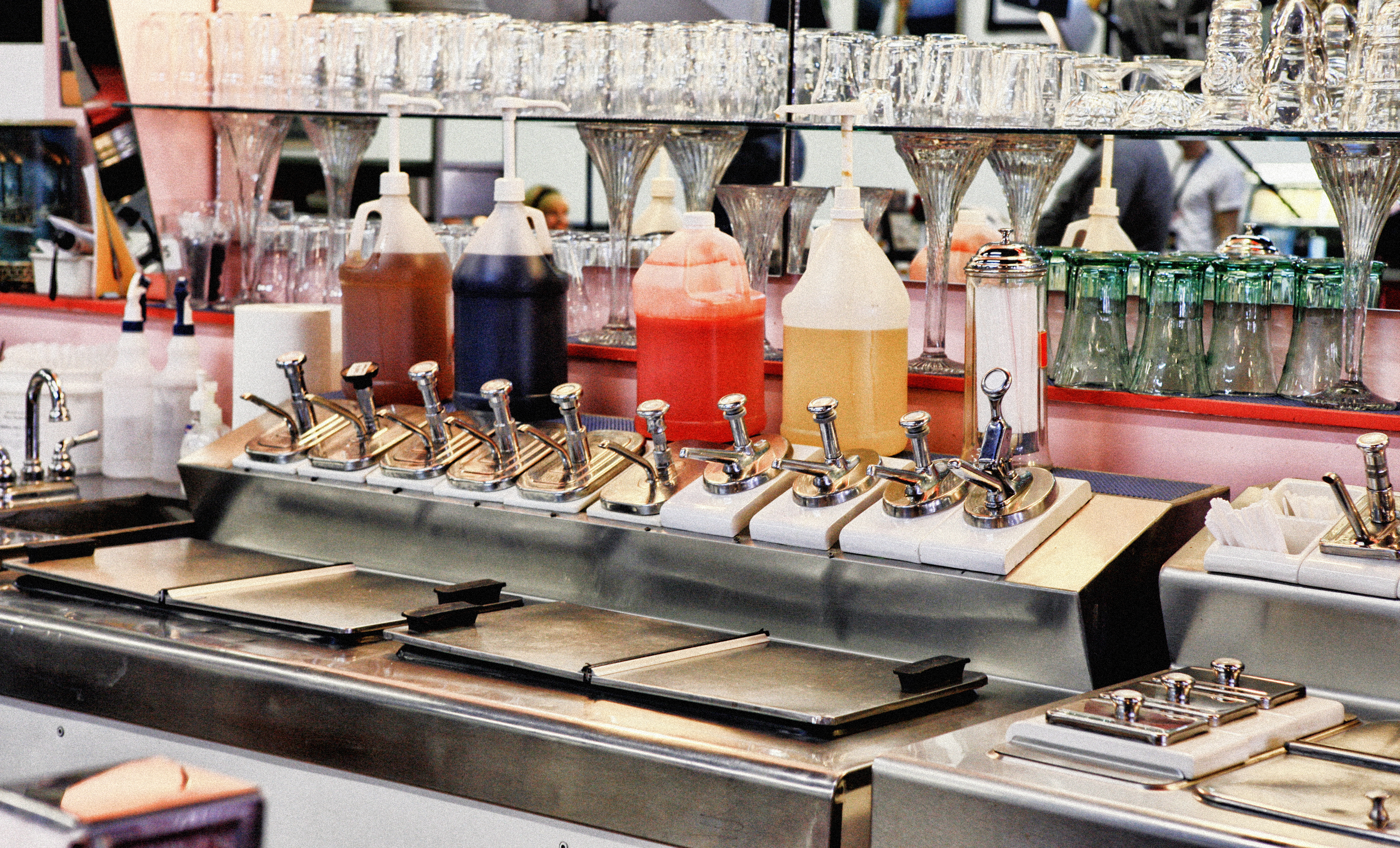 A vintage soda fountain setup with syrup dispensers, milkshake containers, glassware, and other equipment typically found in a retro diner