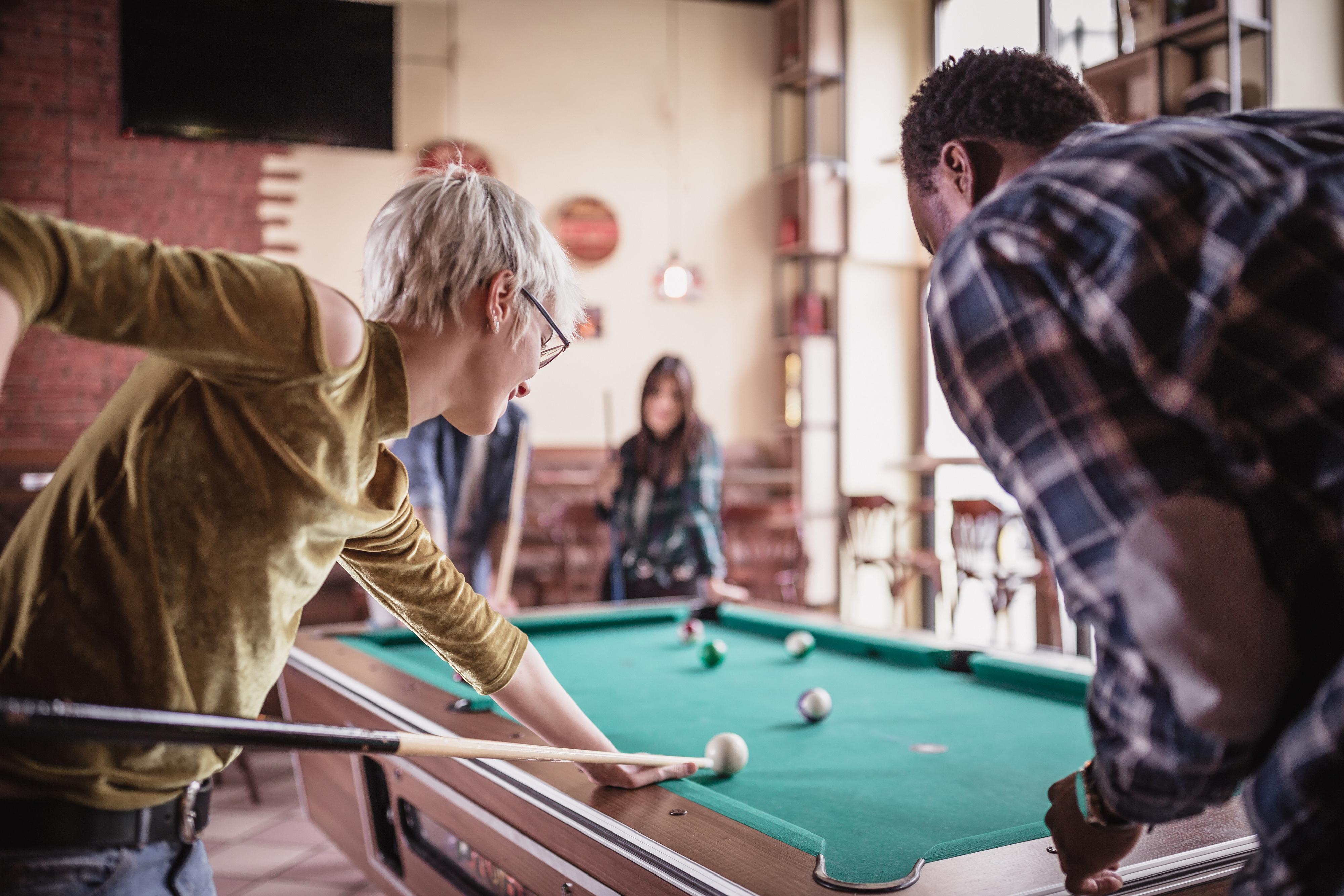 Three people playing pool in a casual indoor setting. One person, with short hair, takes a shot while another in a plaid shirt watches. The third person observes.