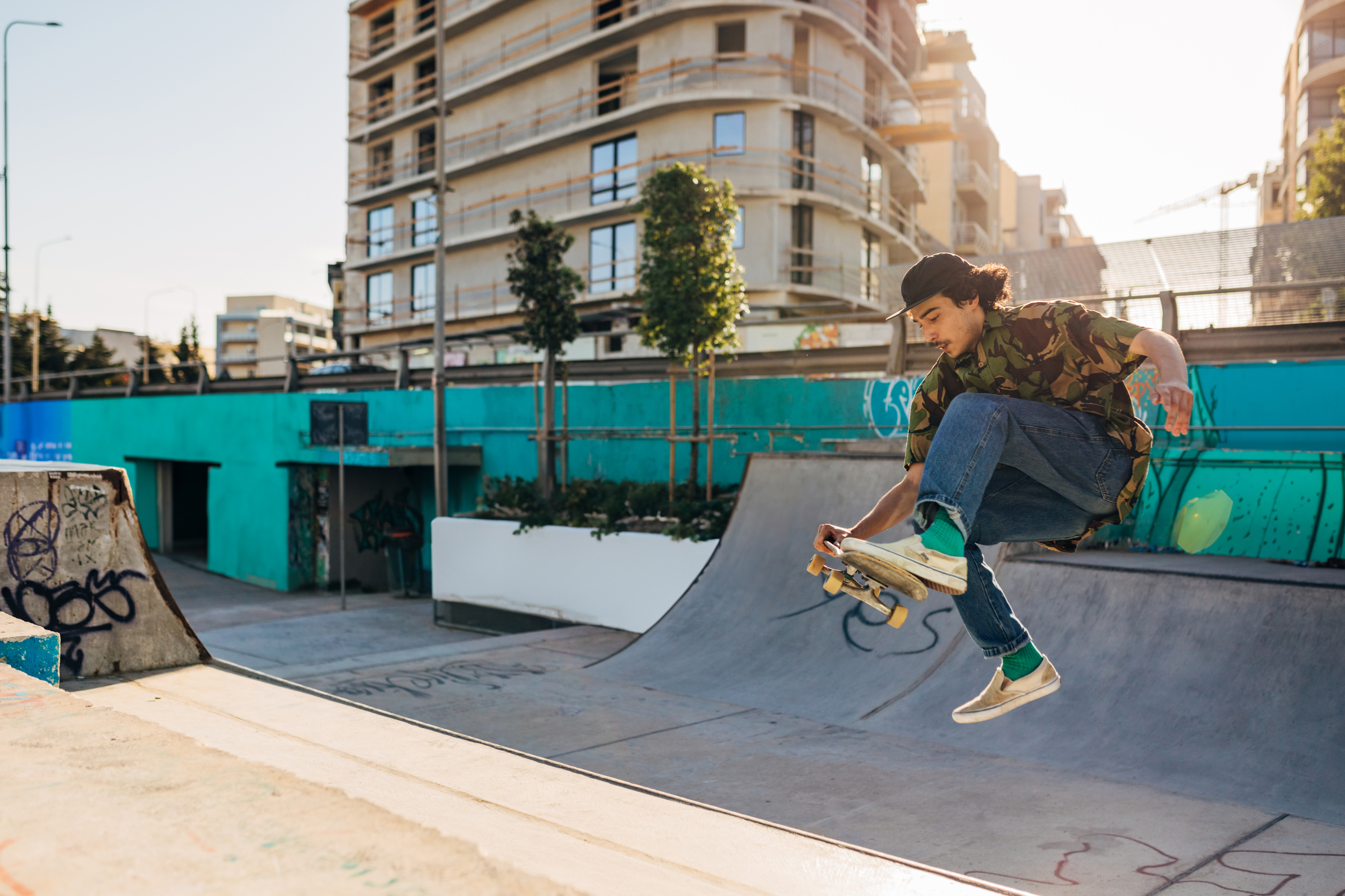 A person skateboards mid-air in a skatepark, performing a trick near a ramp, with urban buildings in the background