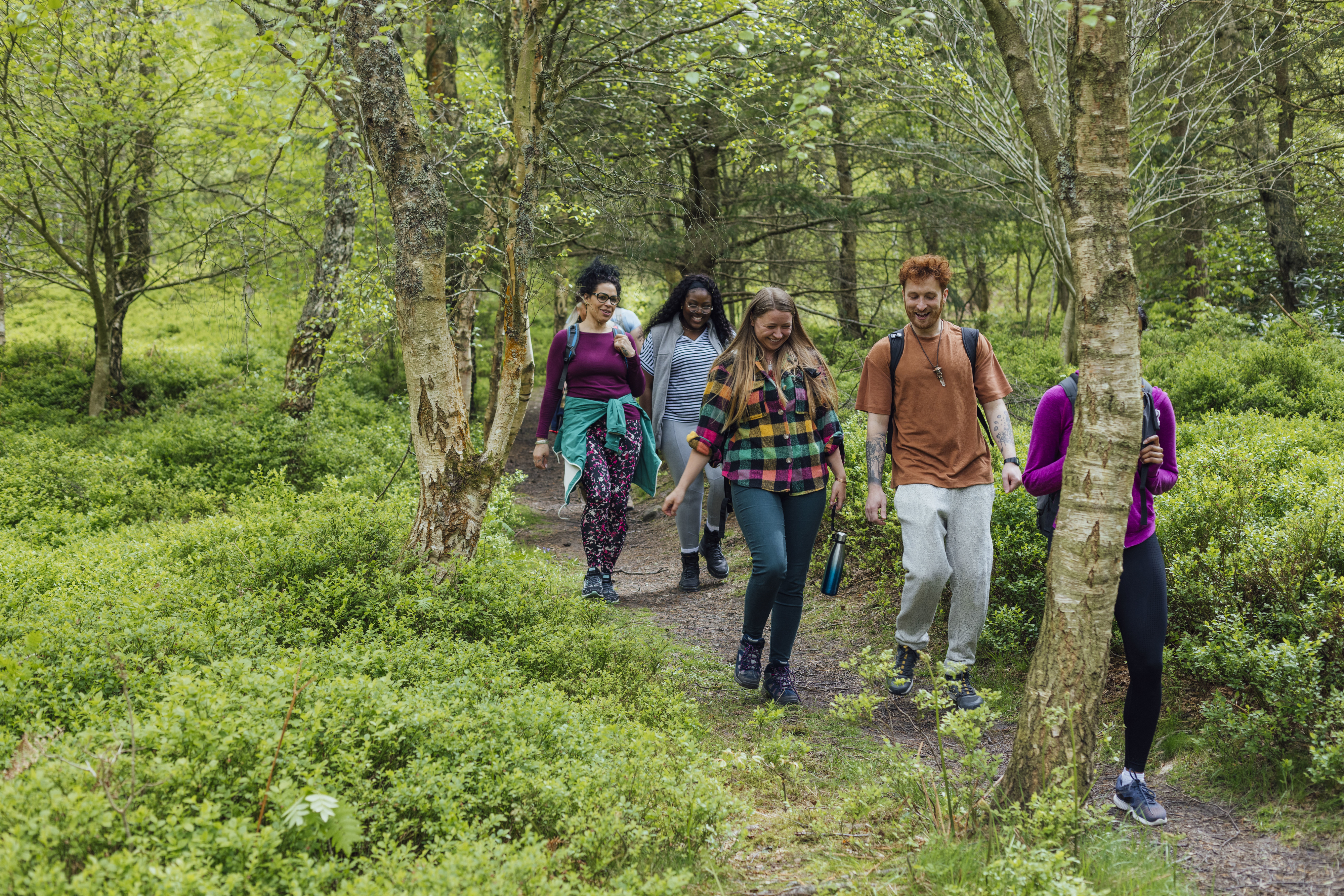 Group of five people, including three women and two men, hiking through a forest. They are smiling and walking on a trail