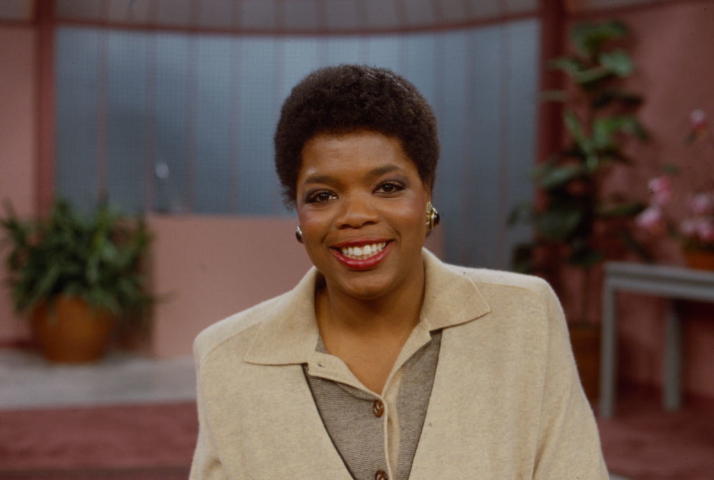 Oprah Winfrey smiles in a studio wearing a beige blazer over a grey top, with plants and curtains in the background