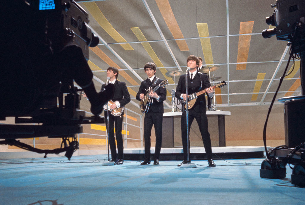 Paul McCartney, George Harrison, and John Lennon perform on a TV stage with instruments, with Ringo Starr on drums in the background