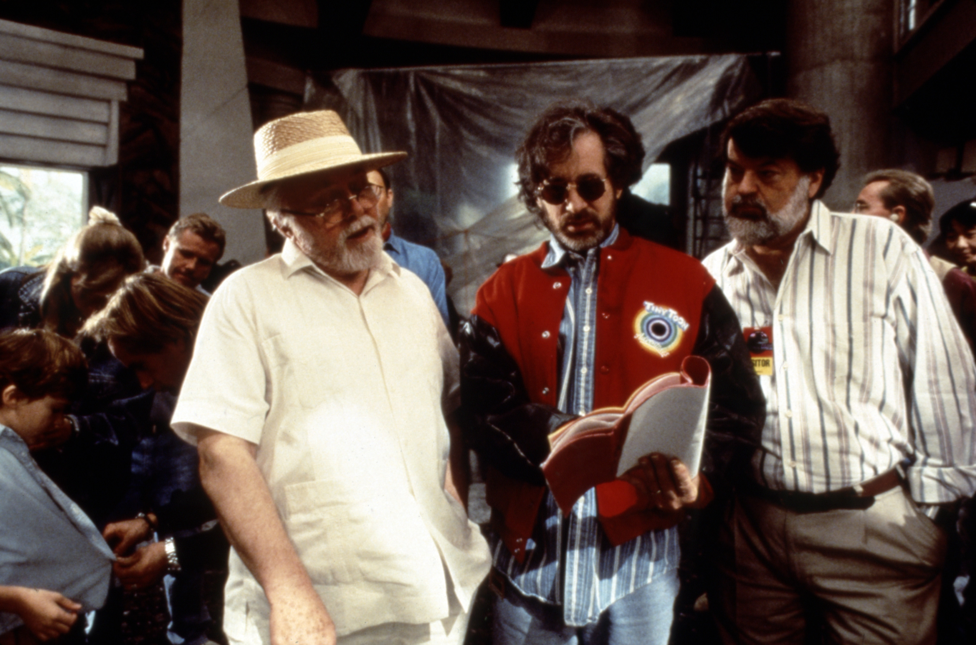 Richard Attenborough, Steven Spielberg in a red jacket, and a unidentified person on a film set. Spielberg is holding a book, likely a script