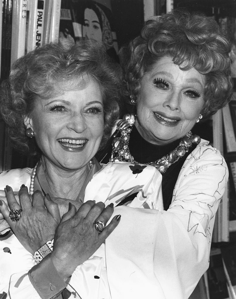 Betty White and Lucille Ball smiling and hugging, wearing elegant dresses adorned with jewelry